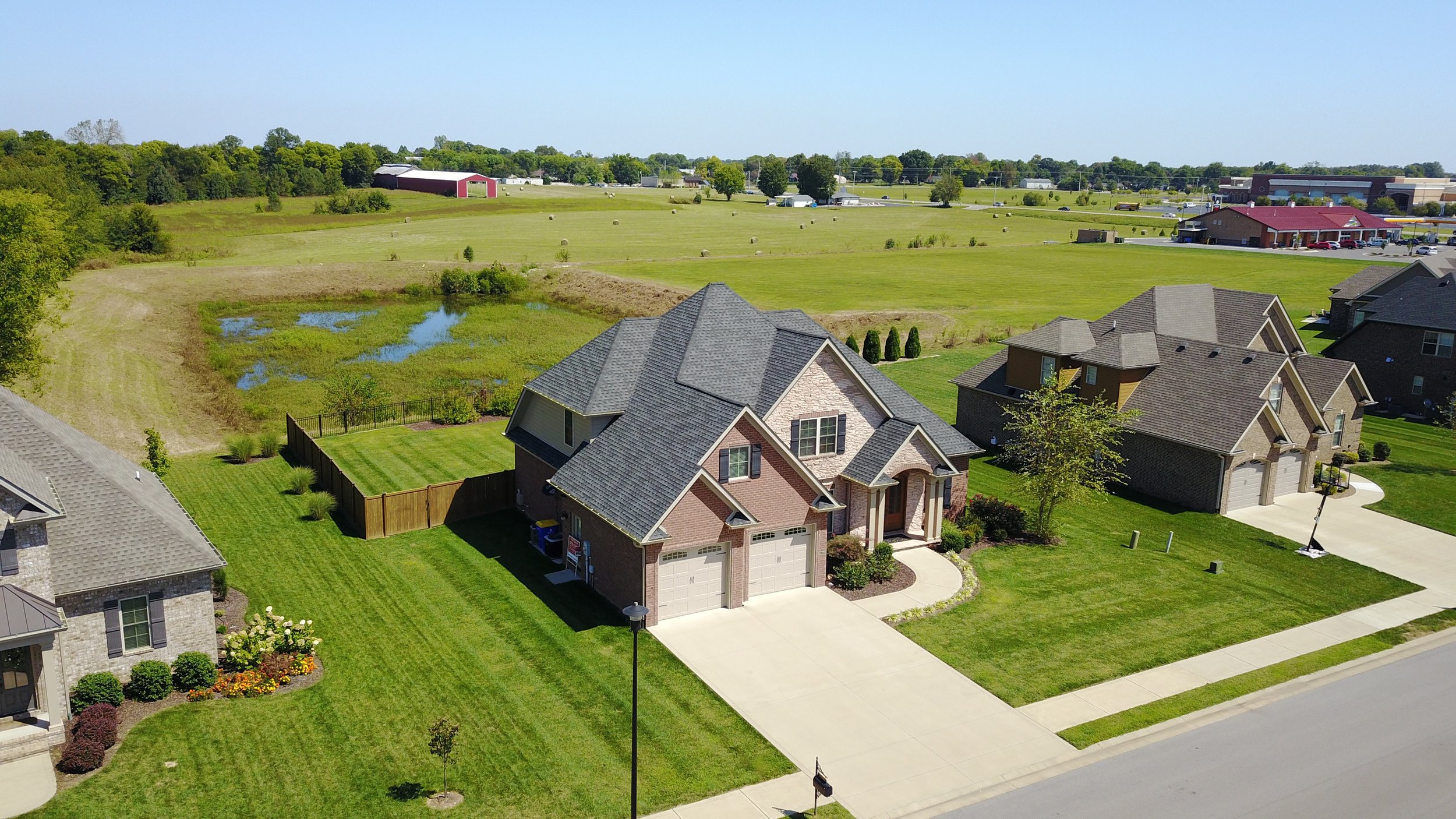 Aerial view of suburban houses with green lawns, driveway, and surrounding open fields.