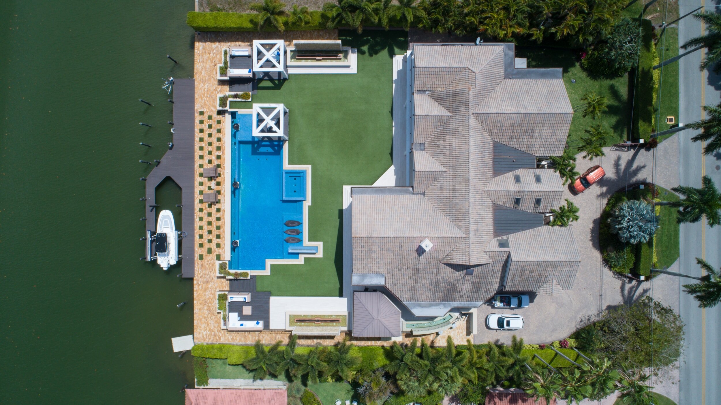 Aerial view of a luxurious waterfront property with a large pool, a docked boat, and a spacious house surrounded by greenery and palm trees. A driveway with parked vehicles is visible, leading to the road.