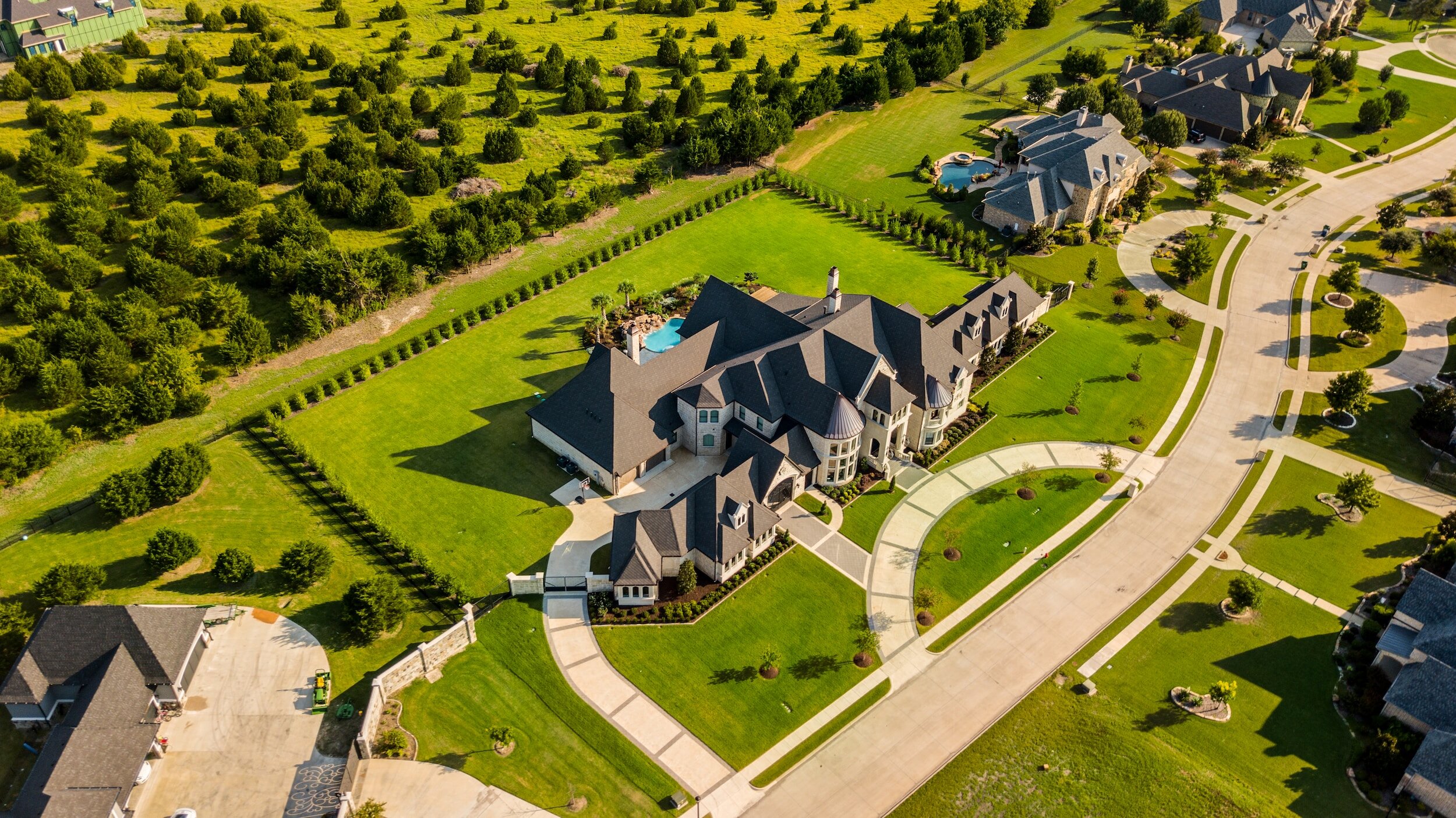 Aerial view of a large mansion with a pool and extensive lawns, surrounded by a residential neighborhood and landscaped greenery.