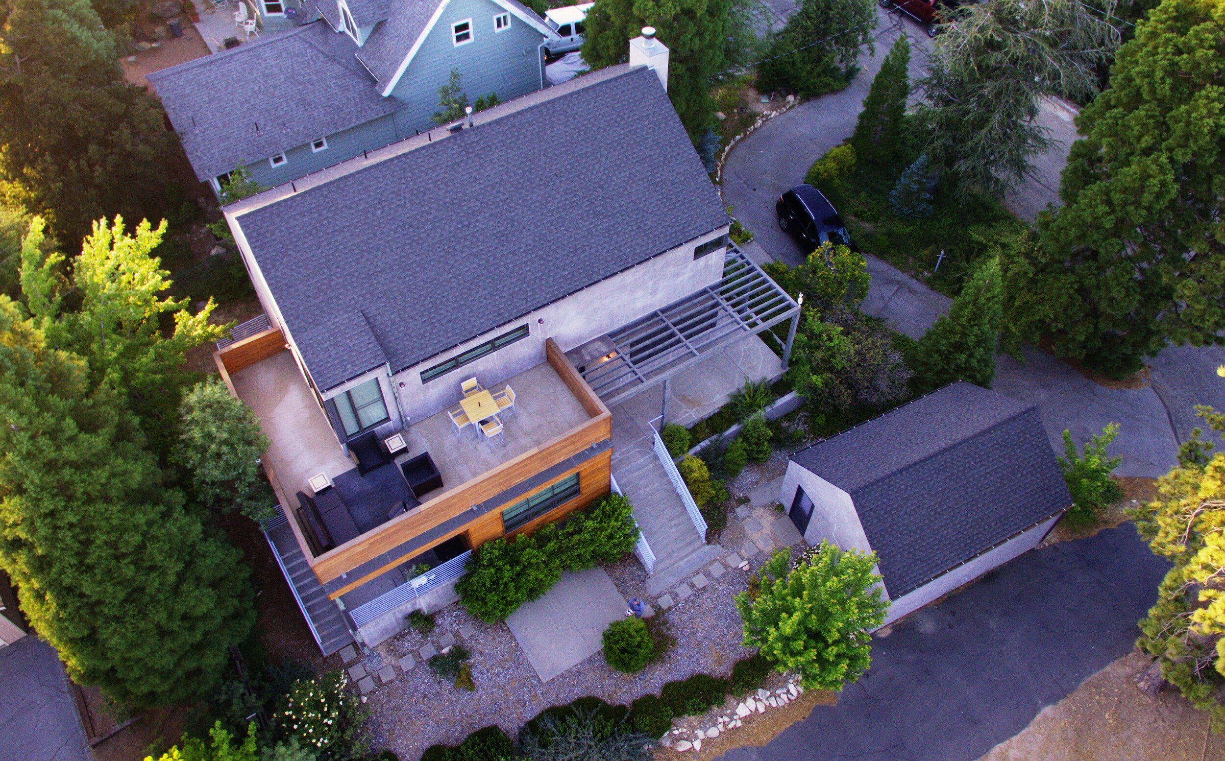 Aerial view of a modern house with a flat roof and a wooden patio area, surrounded by trees and a paved driveway. There are outdoor seating arrangements and a small outbuilding nearby.