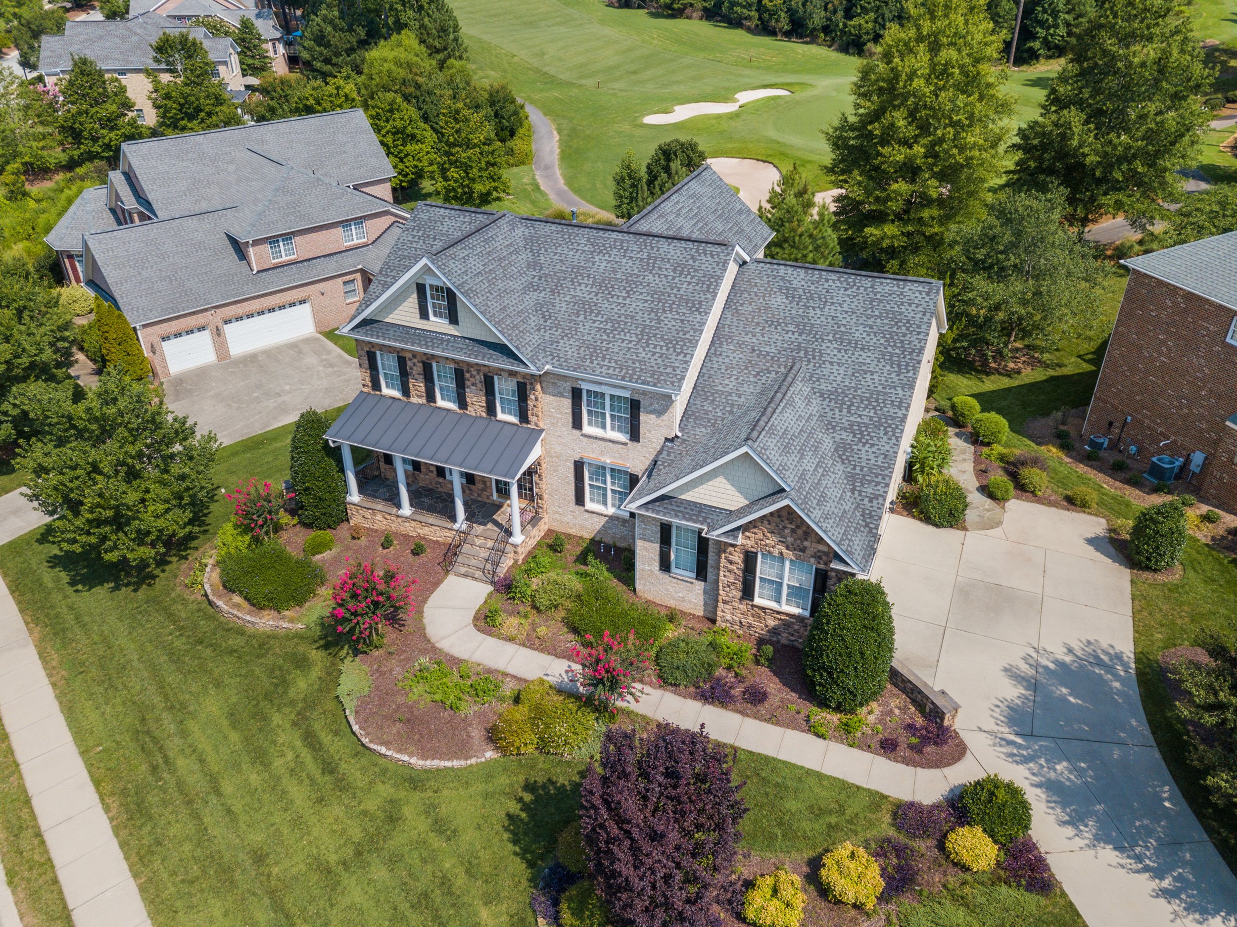 Aerial view of a large suburban house with a landscaped garden, driveway, and a neighboring house. The property includes a well-manicured lawn and is surrounded by trees. A golf course can be seen in the background.