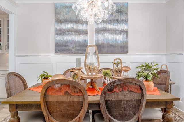 Elegant dining room with wooden table and wicker chairs, orange placemats, potted plants, decorative light fixture, and abstract wall art.