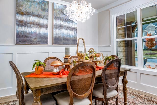 Elegant dining room with a wooden table, cushioned chairs, and orange accents. A modern chandelier hangs overhead, and artwork adorns the walls. Large window offers a view outside.