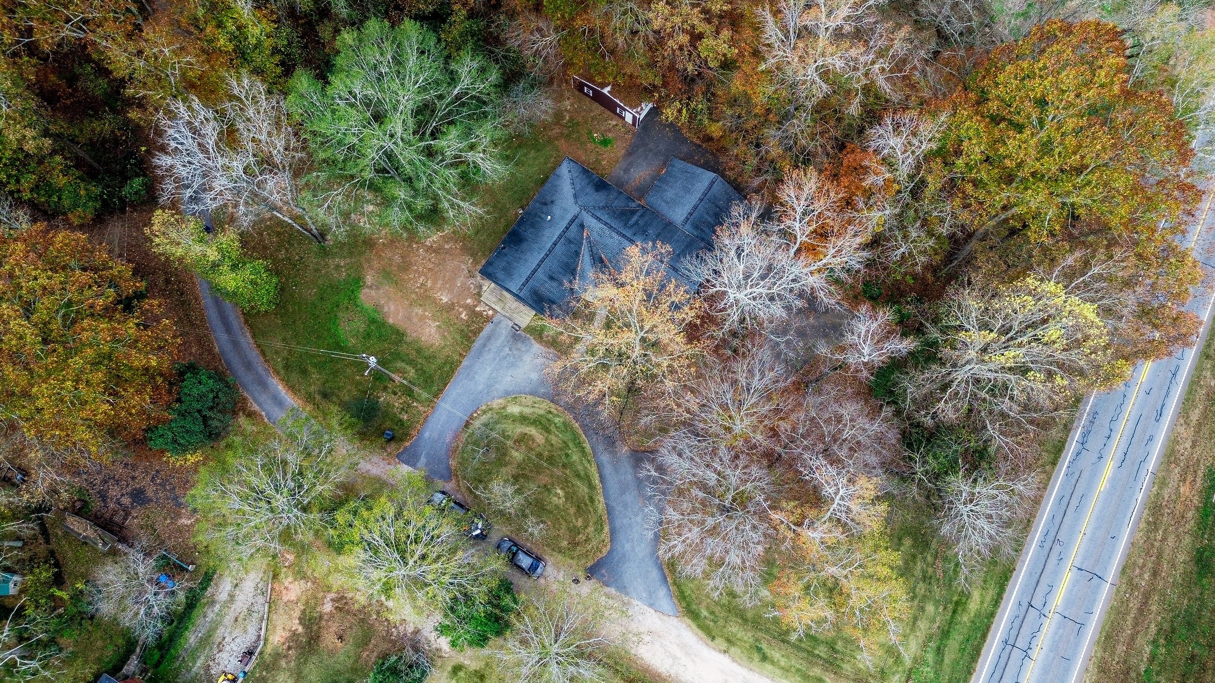 Aerial view of a house surrounded by trees and a driveway leading to a road. The trees have autumn leaves, and there are several cars parked near the house. A paved road is visible to the right.