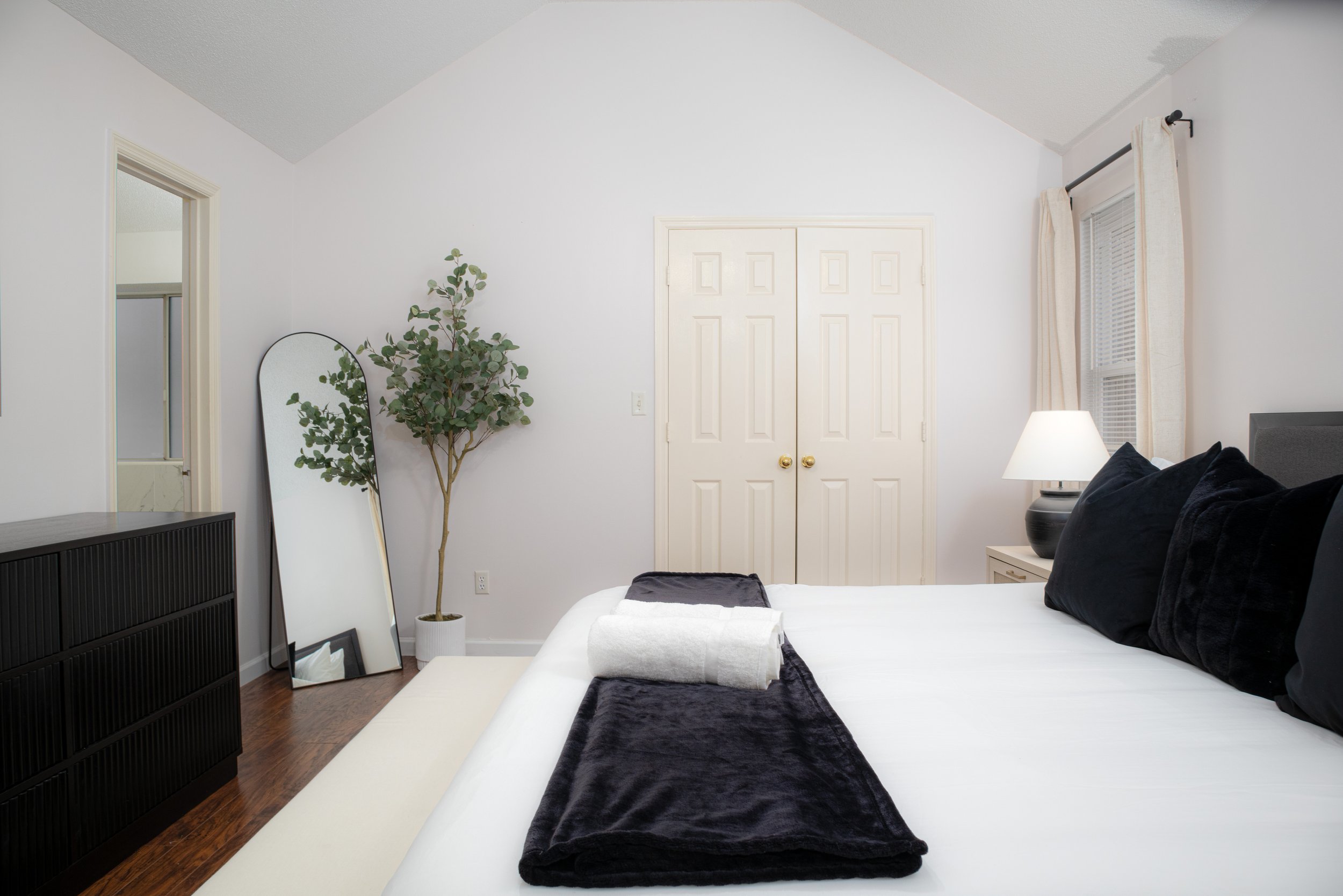 Modern bedroom with white walls, cozy bed with black pillows and blanket, rolled white towels, black dresser, large floor mirror, potted plant, double doors, and a lamp on a bedside table.