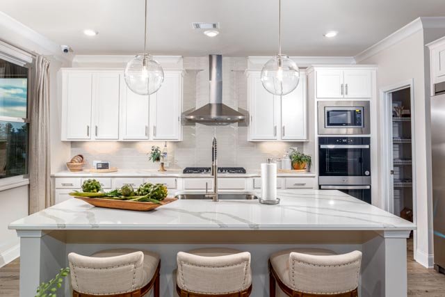 Modern kitchen with white cabinets, stainless steel appliances, and a large central island featuring a white countertop and three upholstered chairs. Hanging pendant lights are above the island, and there are potted plants and decorative items on the