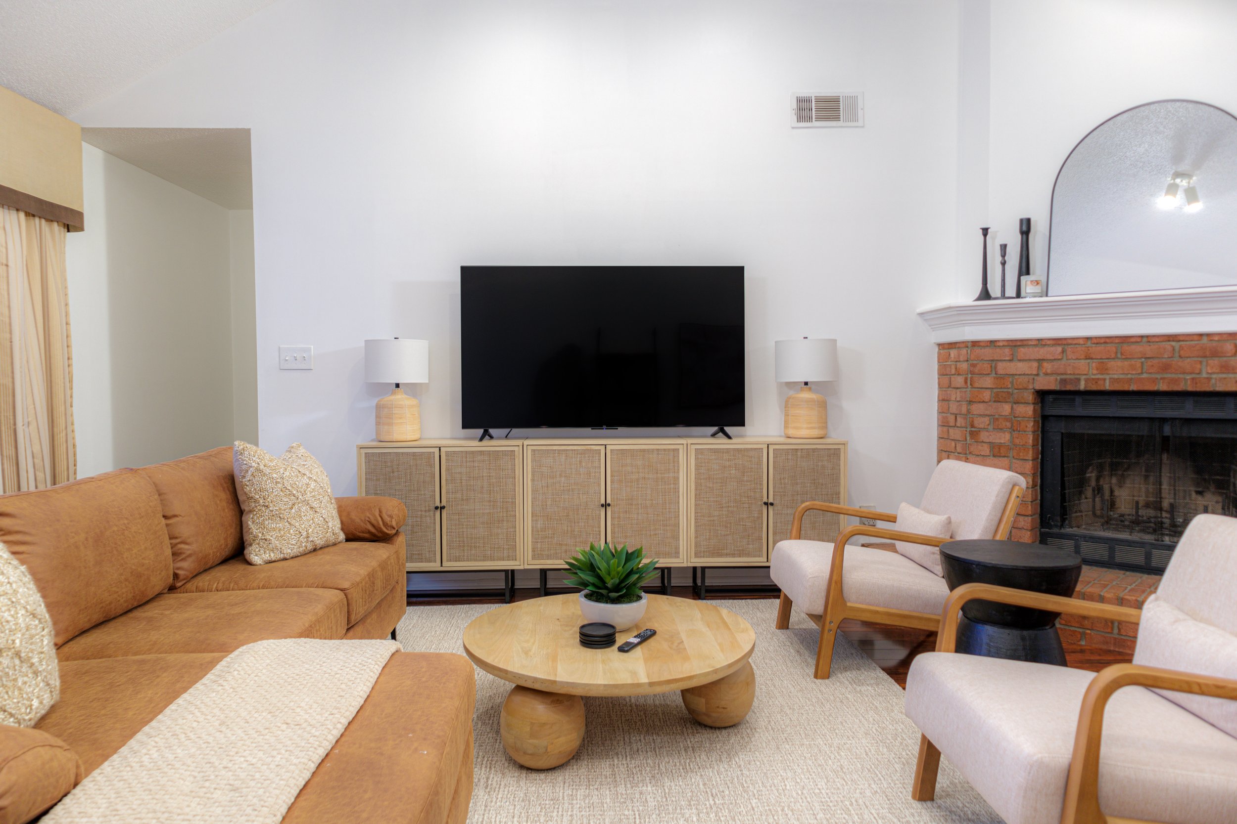 Living room with brown sofa, two beige chairs, wooden coffee table, TV on a console, lamps, brick fireplace, and decorative items.