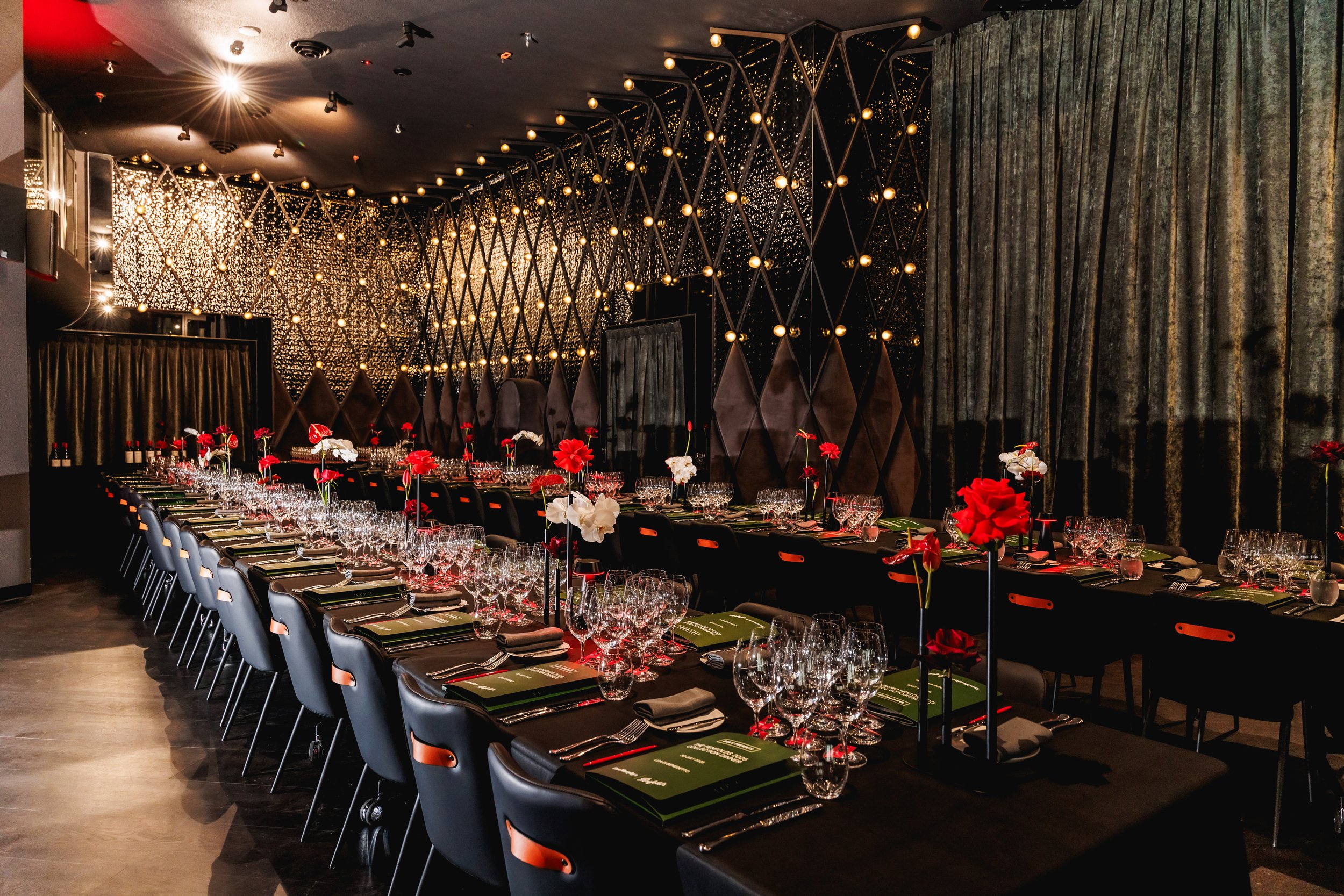 A decorated banquet table set for a formal event in a luxurious, modern indoor venue with black tablecloths, floral centerpieces, glassware, and place settings, illuminated by warm lighting and decorative wall accents.