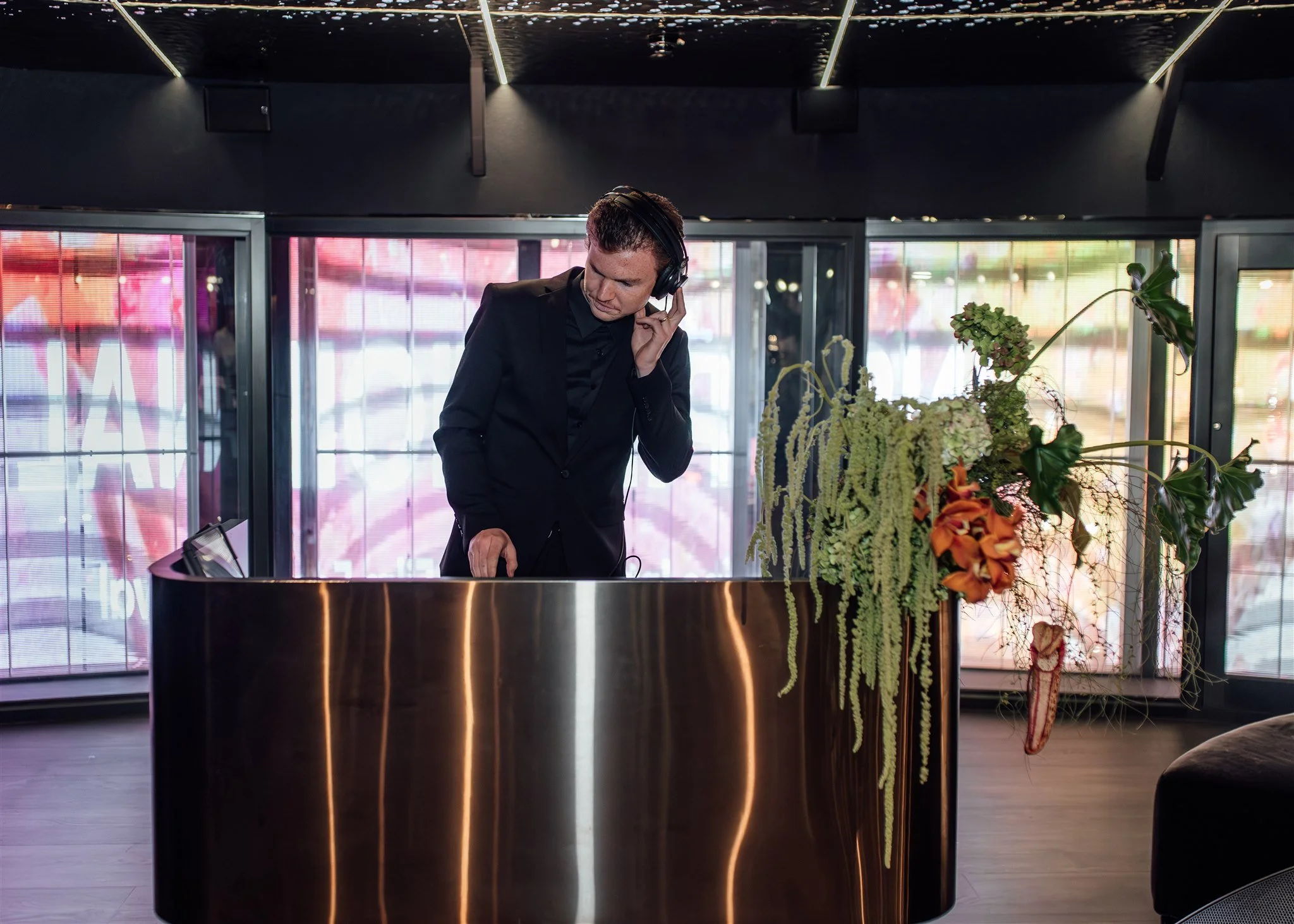A man in a black suit with headphones stands behind a curved, reflective metallic bar in a modern, dimly lit room. Large windows with colorful, blurred city lights are in the background, and there is a floral arrangement with green, orange, and purpl