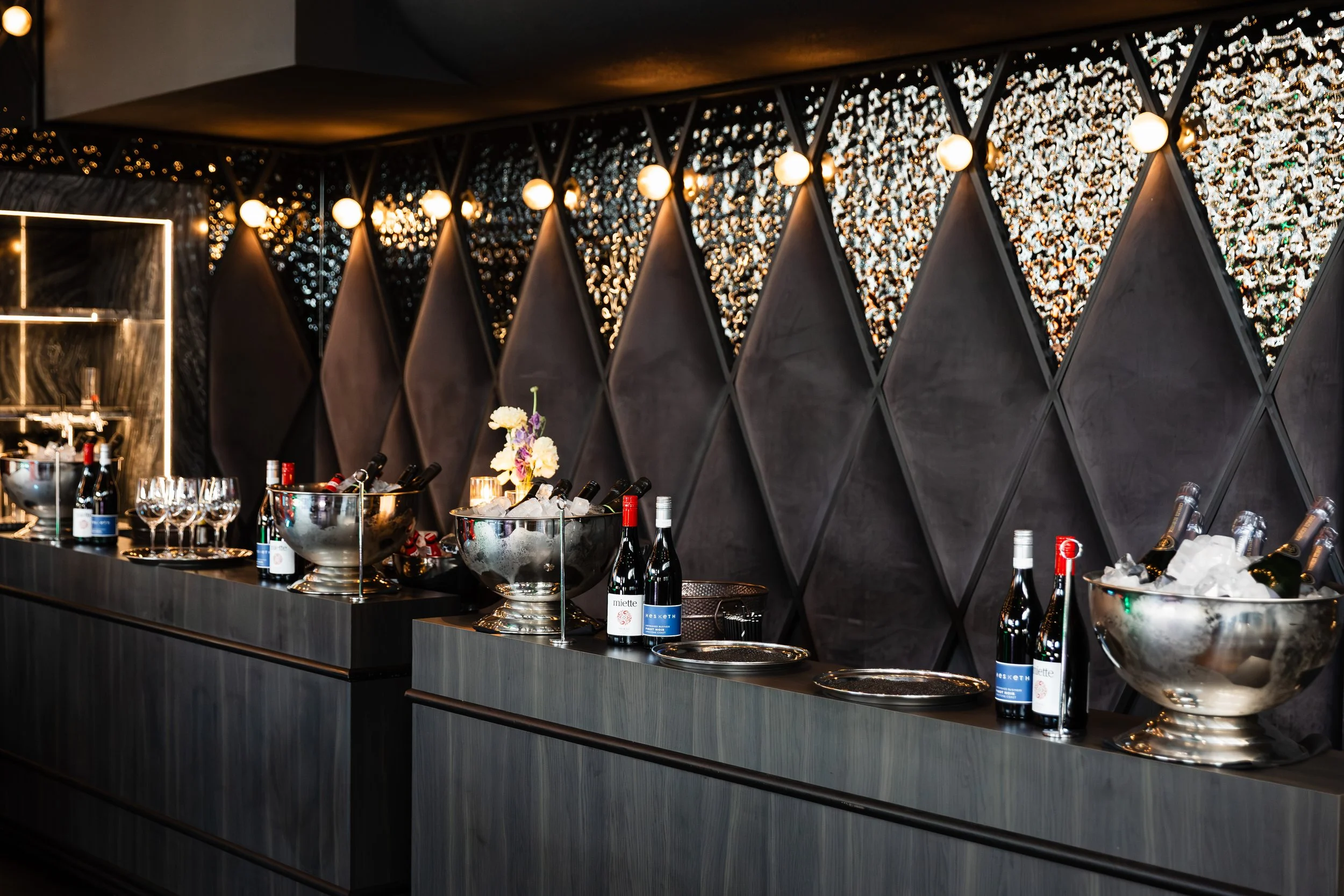 Bar area with champagne bottles, ice buckets, and glasses, featuring decorative wall with geometric patterns and string lights.
