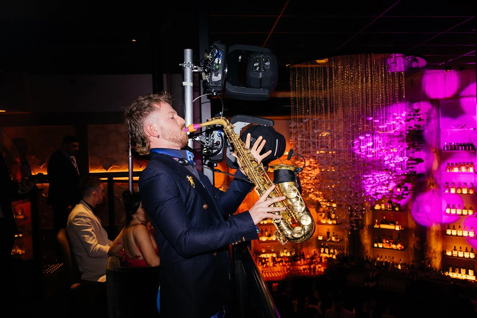 A saxophone player performing at a bar or nightclub with vibrant purple and orange lighting.