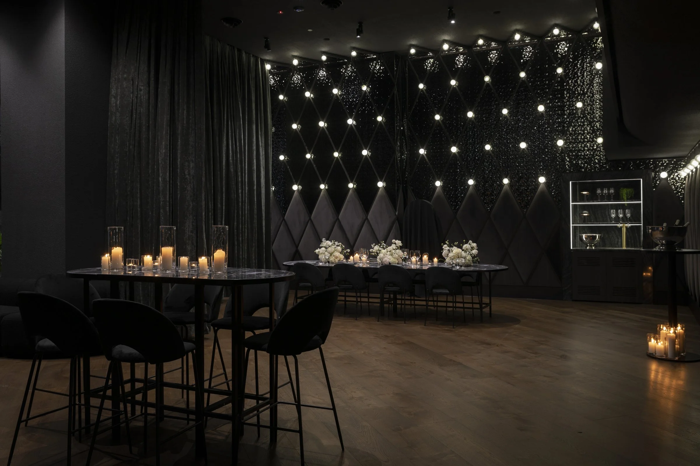 Elegant dimly lit event space with candles on tables and floral arrangements, featuring black walls and modern lighting fixtures.