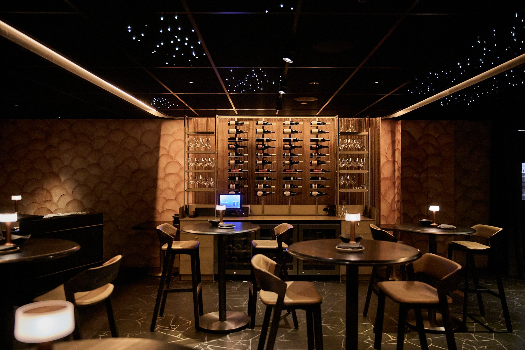 Elegant restaurant interior with round tables, chairs, wine bottle display on the wall, wine glasses, and soft lighting.