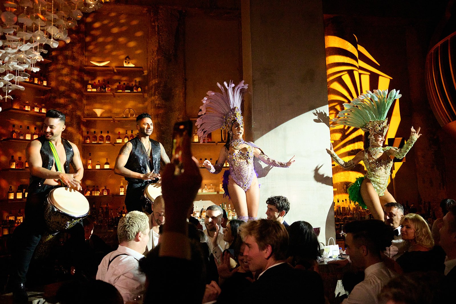 Two performers with elaborate feathered headdresses dance on stage, while two drummers in black vests and no shirts play for an audience in a dimly lit venue with bottles on shelves in the background.
