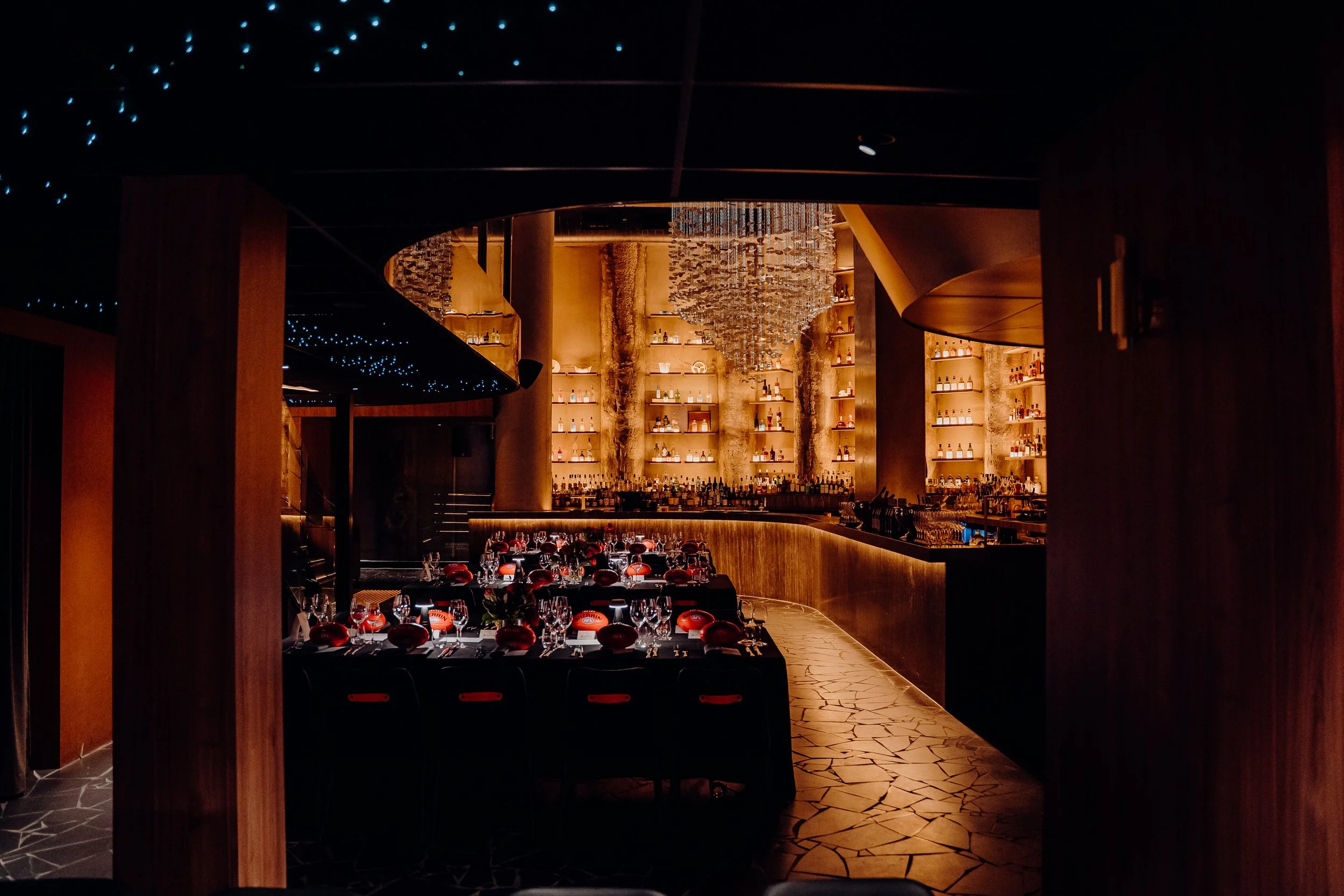 Elegant dimly lit restaurant interior with a bar, shelves displaying bottles, and a chandelier hanging from the ceiling.