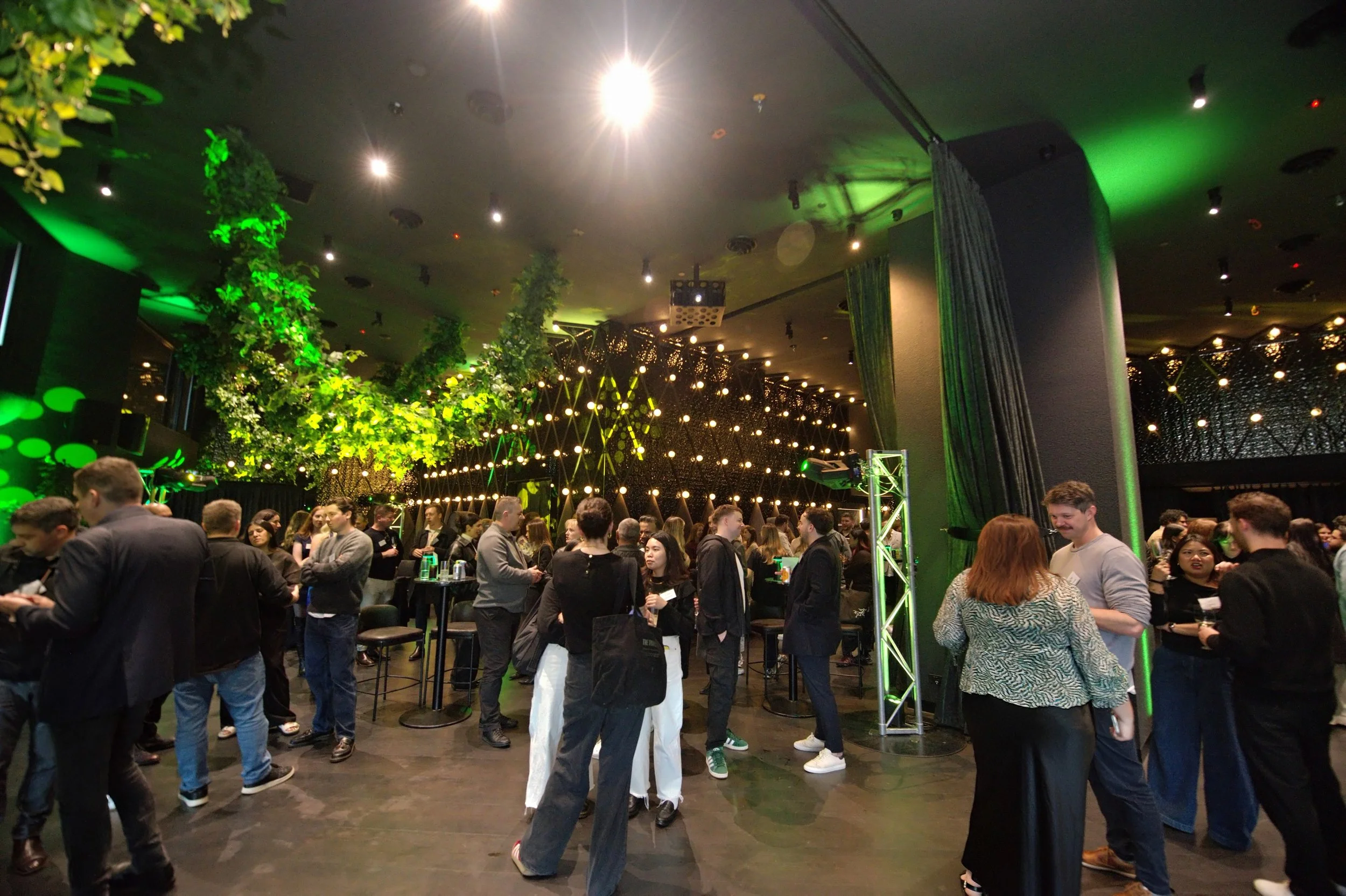 People socializing at a lively indoor event with green lighting, decorative plants, and modern lighting fixtures.