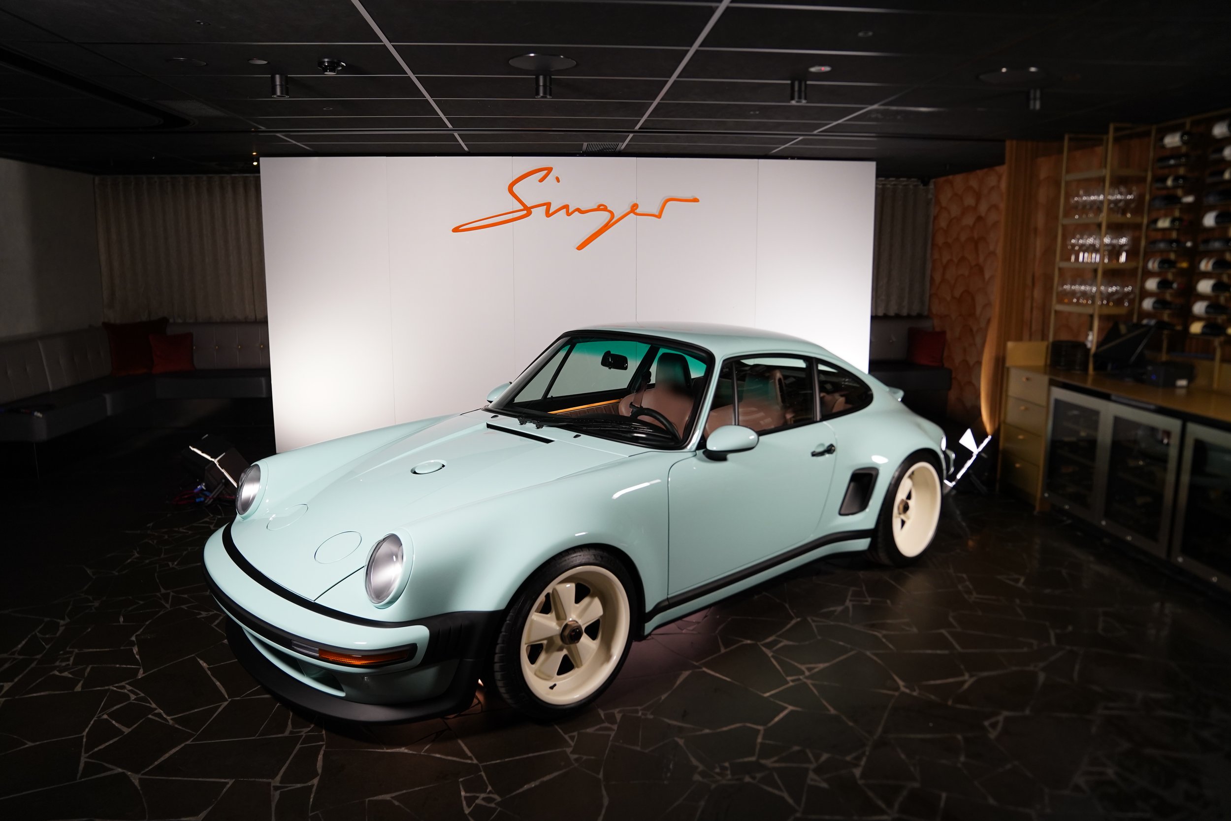 A light green vintage Porsche 911 sports car displayed indoors, with a white wall behind it featuring an orange signature-like logo.