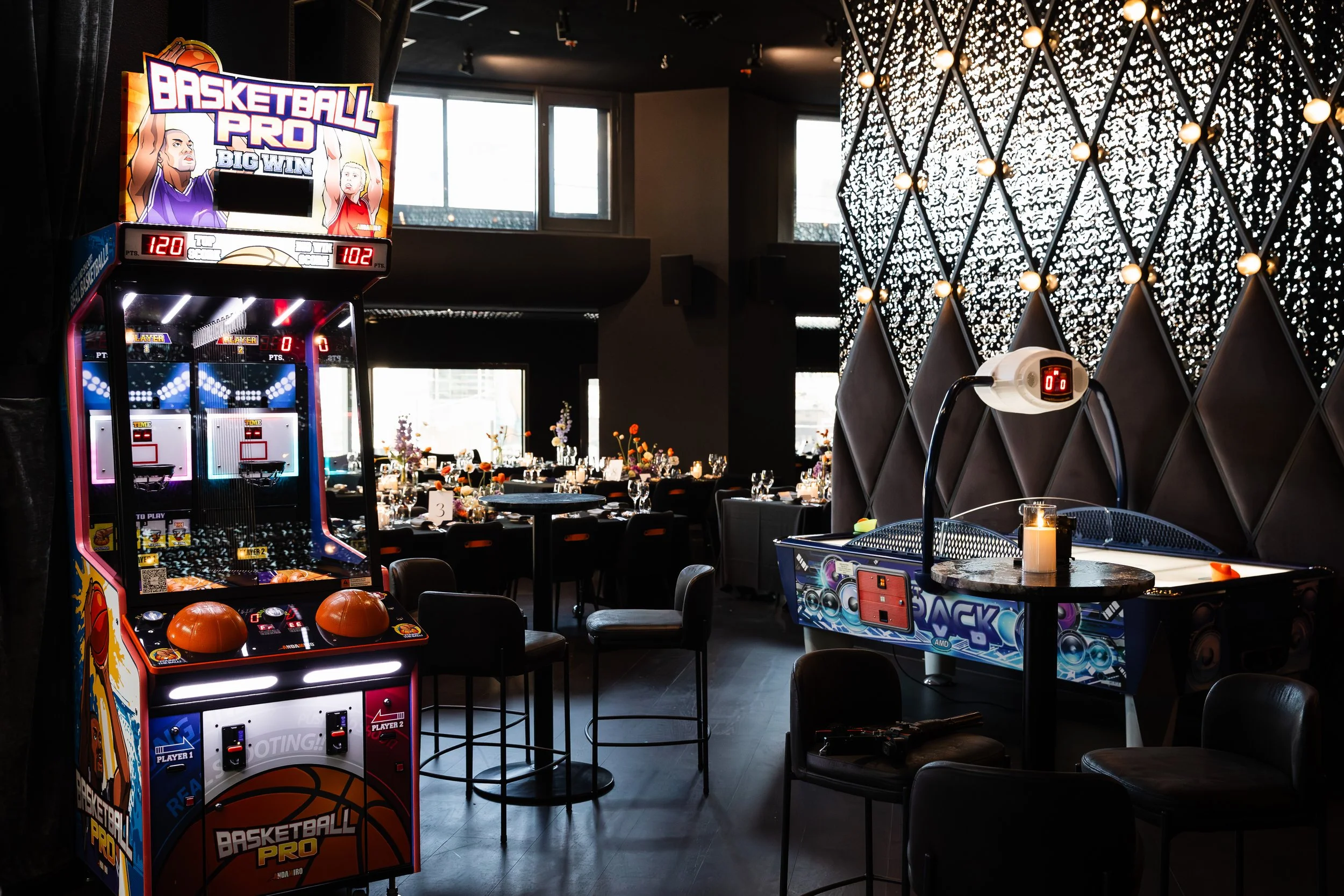An indoor arcade and game room with a basketball arcade game, a tabletop game, and a dining area with tables and chairs decorated for an event.