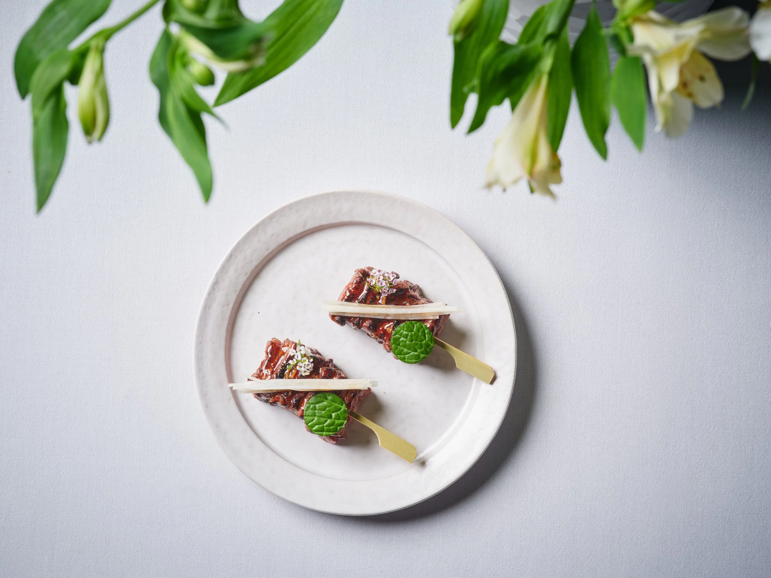 Two gourmet appetizers on a white ceramic plate, garnished with green leaves and white flowers, placed on a light gray surface with green plants in the background.