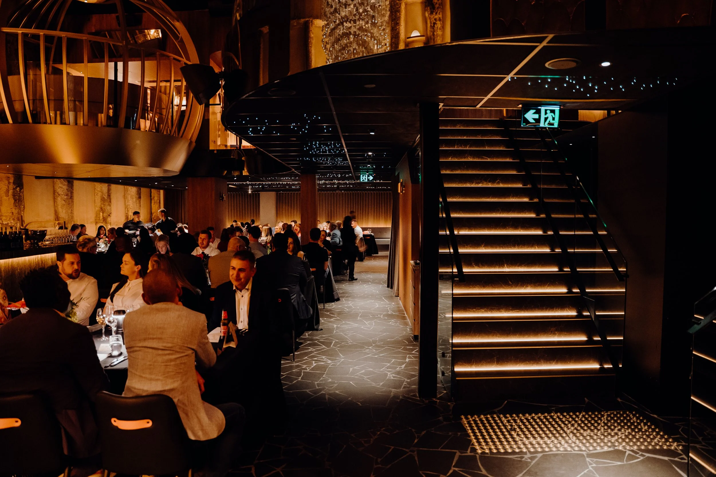 A bustling upscale restaurant interior with groups of people dining at black tables, warm lighting, wooden accents, and a staircase with illuminated steps.