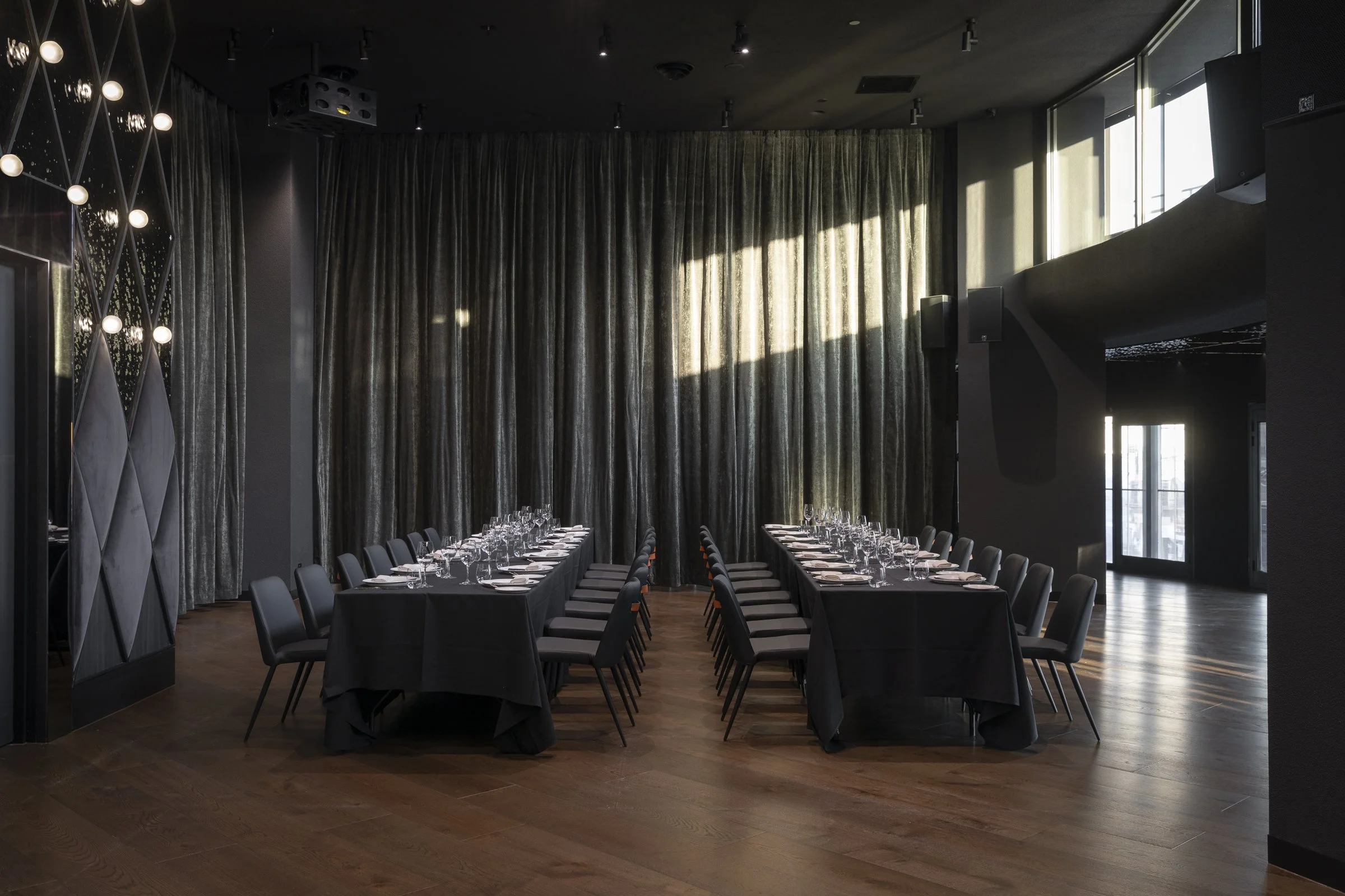 An elegant dining room with two long black tables set with white plates, glasses, and silverware, surrounded by black chairs, and large curtains covering a window with sunlight streaming in.