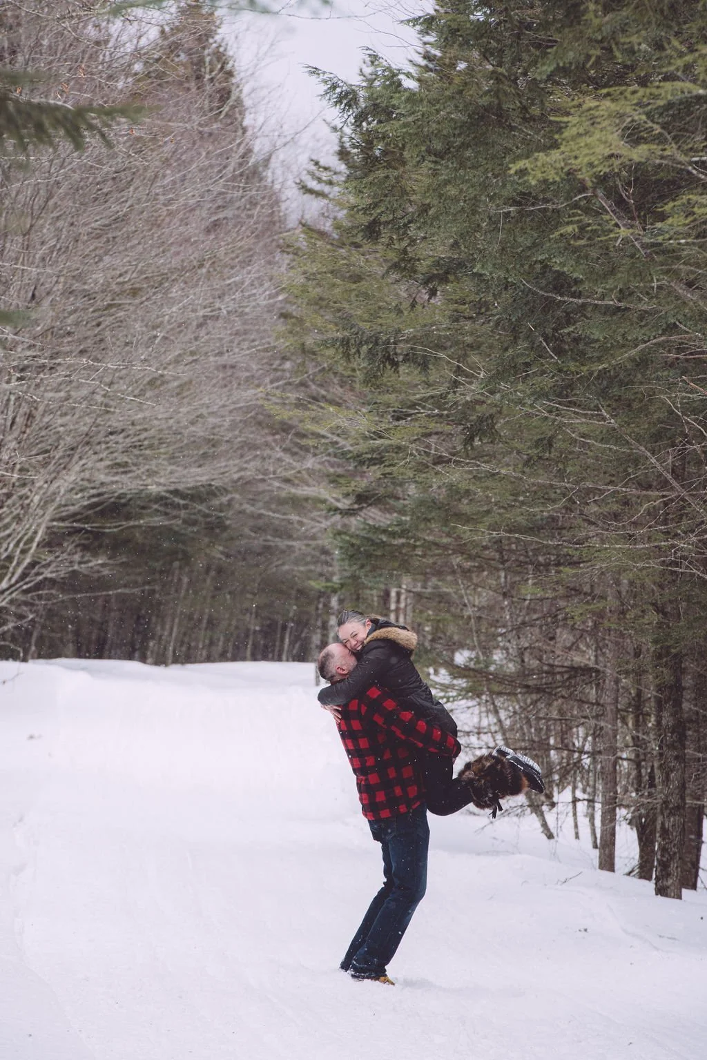 Victoria is being lifted up be her fiancé with a joyous smile while it was snowing during their winter engagement session at The Basin in Franconia Notch, NH. Photography by Mike Indi.
