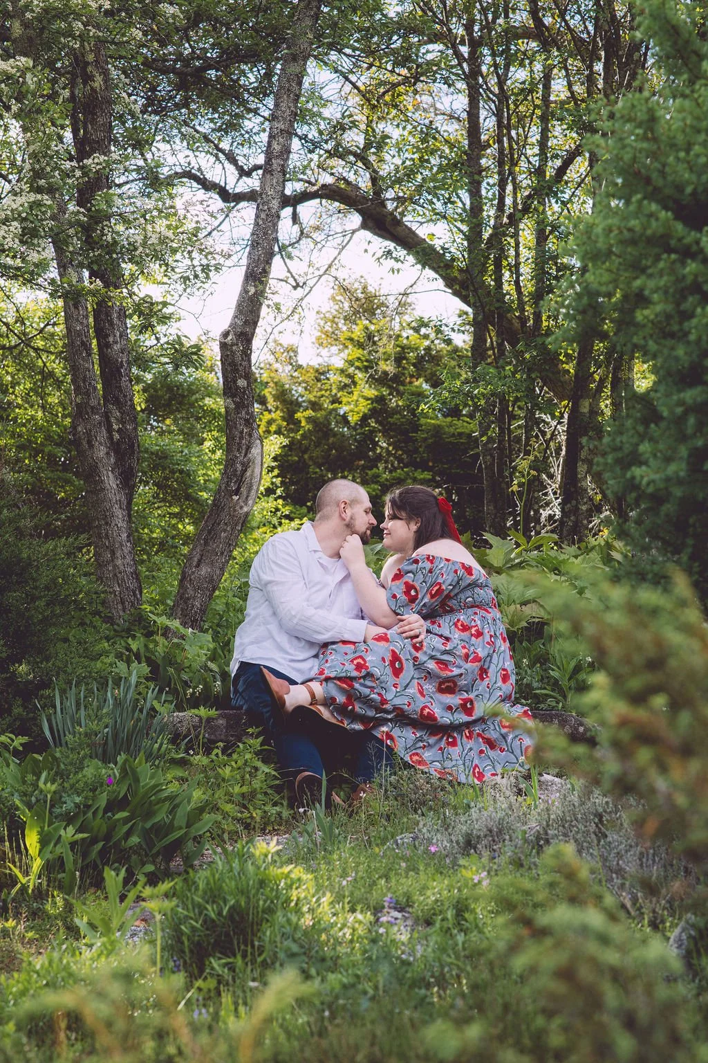 Kelly and Patrick leaning in for a passionate kiss while sitting on a stone bench in a lush garden during their engagement session at the John Hay Estate at The Fells in Newbury, NH. Photography by Mike Indi.