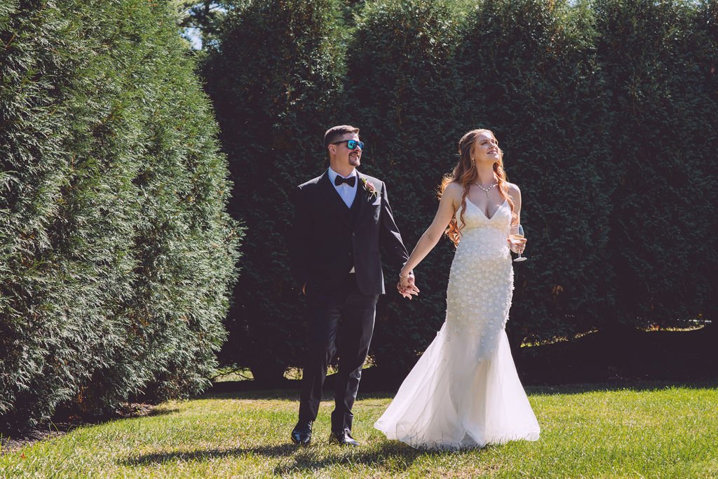 A bride holding a glass of white wine and holding handing with her groom walk through the hedges at Searles Chapel in Windham, NH during the portrait session on their wedding day. Wedding photography by Mike Indi.