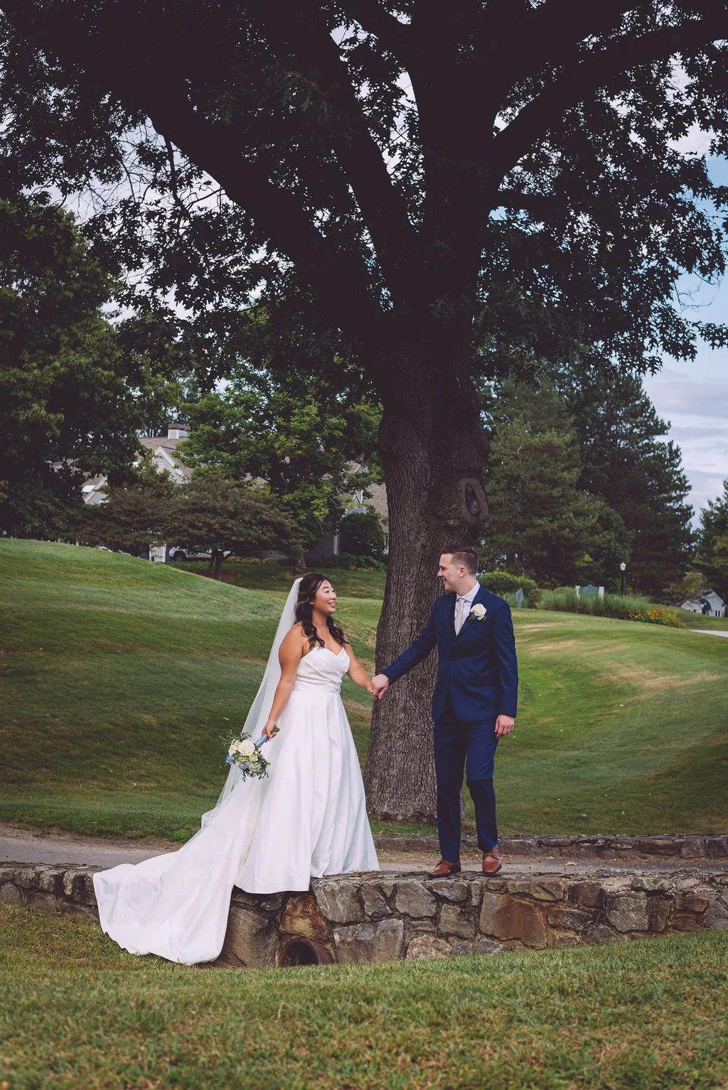 A beautiful mixed-race bride and groom holding hand while walking along a stone wall with gorgeous greenery surrounding them during their wedding at Sky Meadow Country Club in Nashua, New Hampshire. Photography by Mike Indi.