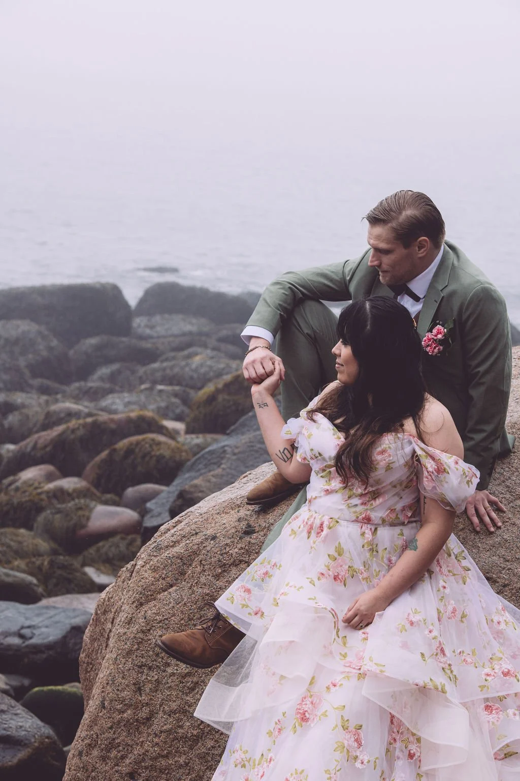 Chelsea and Ryan holding hands and sharing an intimate moment while sitting on a large rock on Hunter's Beach, surrounded by fog, during their adventure elopement in Acadia Nation Park in Bar Harbor Maine. Photography by Mike Indi.