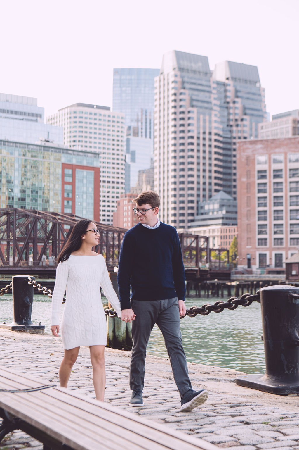 Couple walking hand in hand at Fan Pier Park in Boston's Seaport District with the city skyline behind them during a spring engagement session in Boston, Massachusetts — photography by Mike Indi