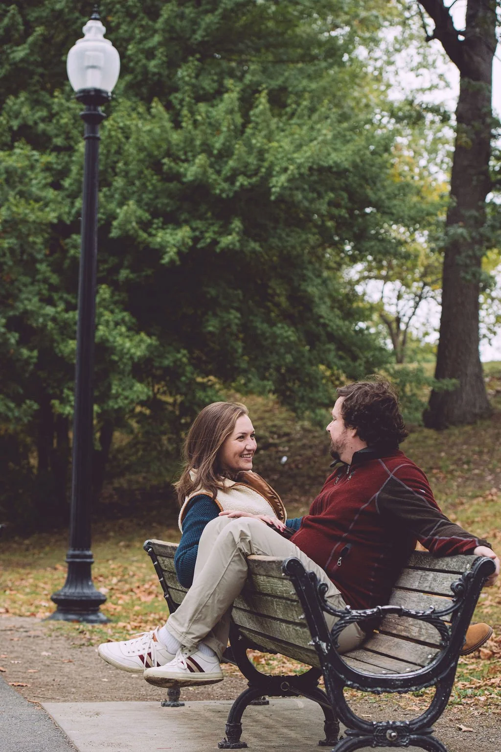 A happy couple laughing together while sitting on the strange U-shaped Hincman's Bench near Jamaica Pond in Boston during their fall engagement session. Engagement photography by Mike Indi.