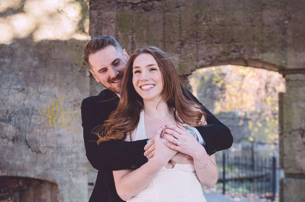 A happy bride smiling while being hugged by her groom during their engagement session at Bancroft's Castle in Groton, MA. Engagement photography by Mike Indi.