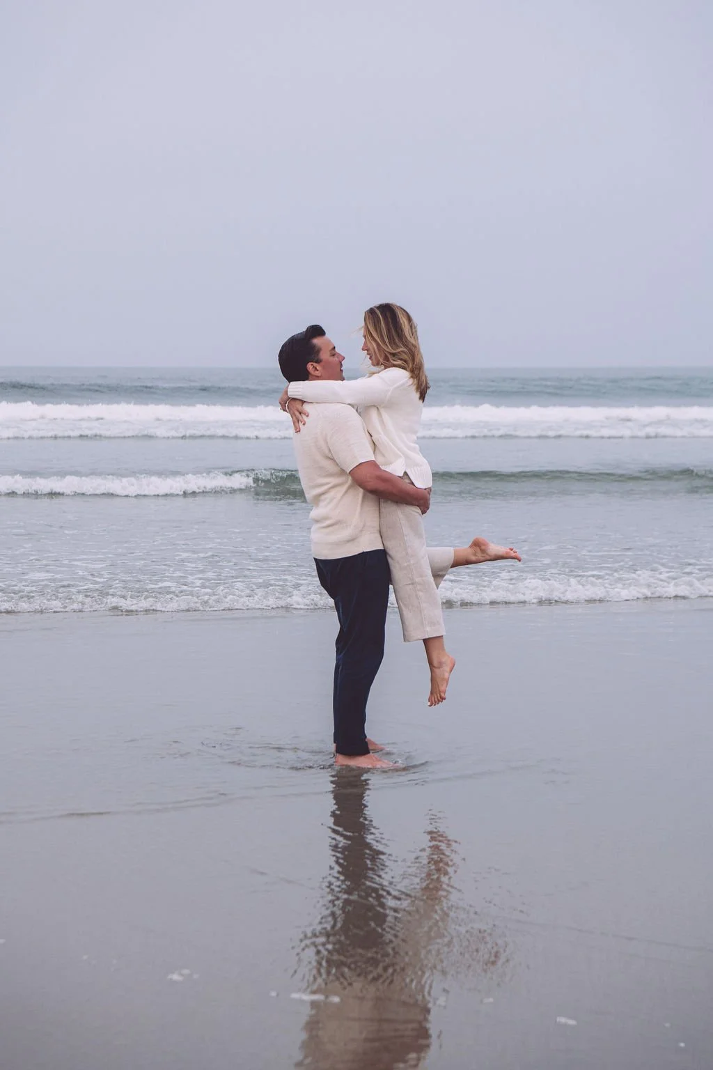 Christopher lifting his fiancé Amanda and spinning her around at the ocean's edge and their reflection can be seen in the water in the foreground on a beach during their early summer engagement session in  Ogunquit, ME. Photography by Mike Indi.