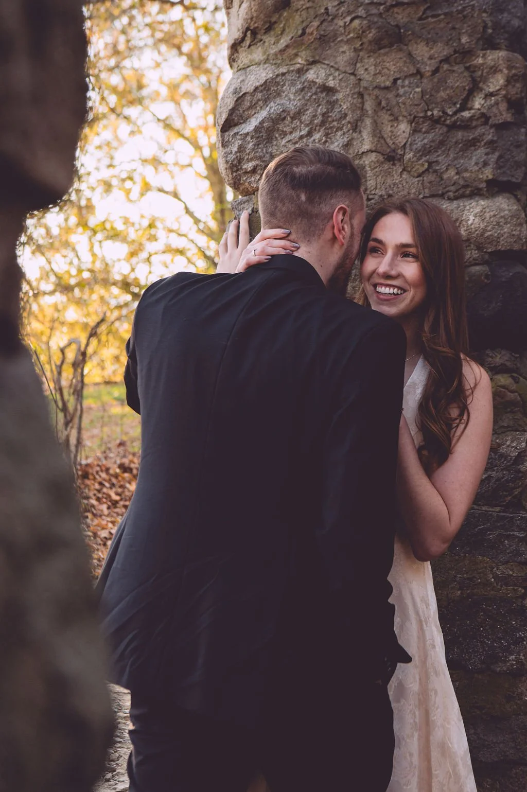 Groom leaning toward his bride against the stone ruins of Bancroft's Castle in Groton, Massachusetts during a fall engagement session — photography by Mike Indi