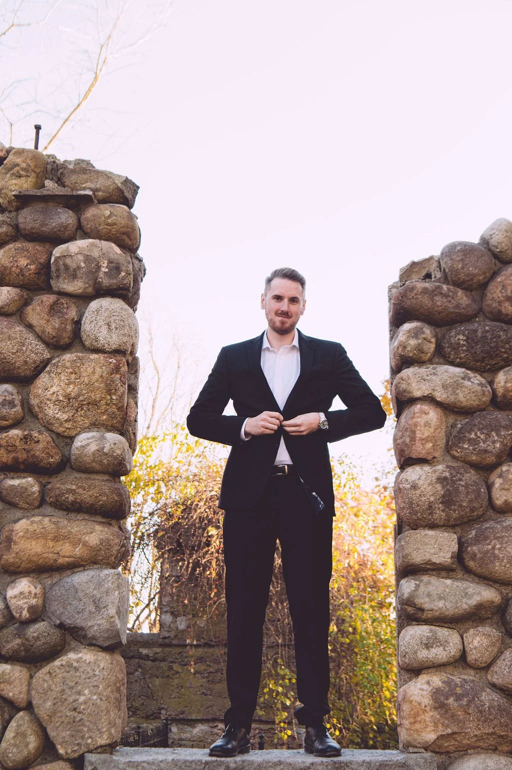 A groom buttoning his suit jacket while standing on a stone castle ruins during his engagement session at Bancroft's Castle in Groton, MA. Engagement photography by Mike Indi.