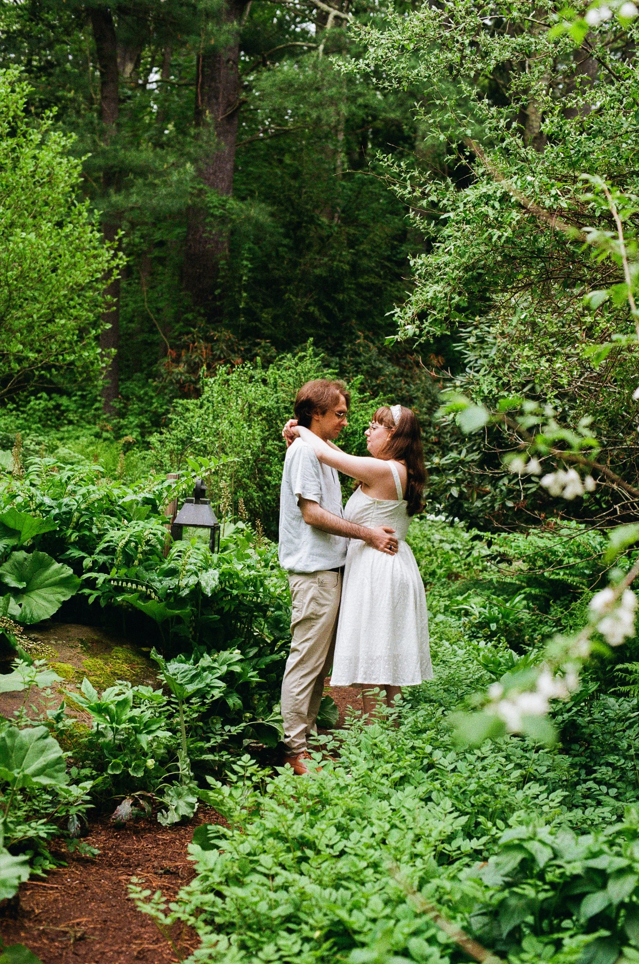 Morgan and Joey in a romantic embrace surround by lush greenery during their spring engagement session at Tarbin Gardens in Franklin, NH - Photography by Mike Indi