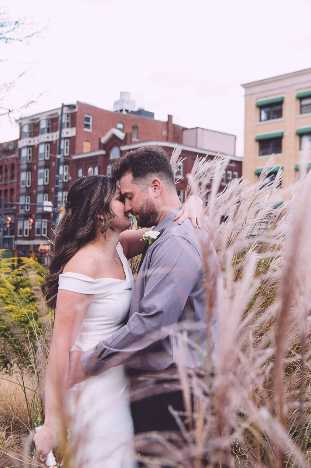 Cassidy and Brandon in a passionate embrace with a mix of bushes and wild flowers with the buildings of downtown Manchester, NH peaking out in the background after their elopement at city hall. Photography by Mike Indi.