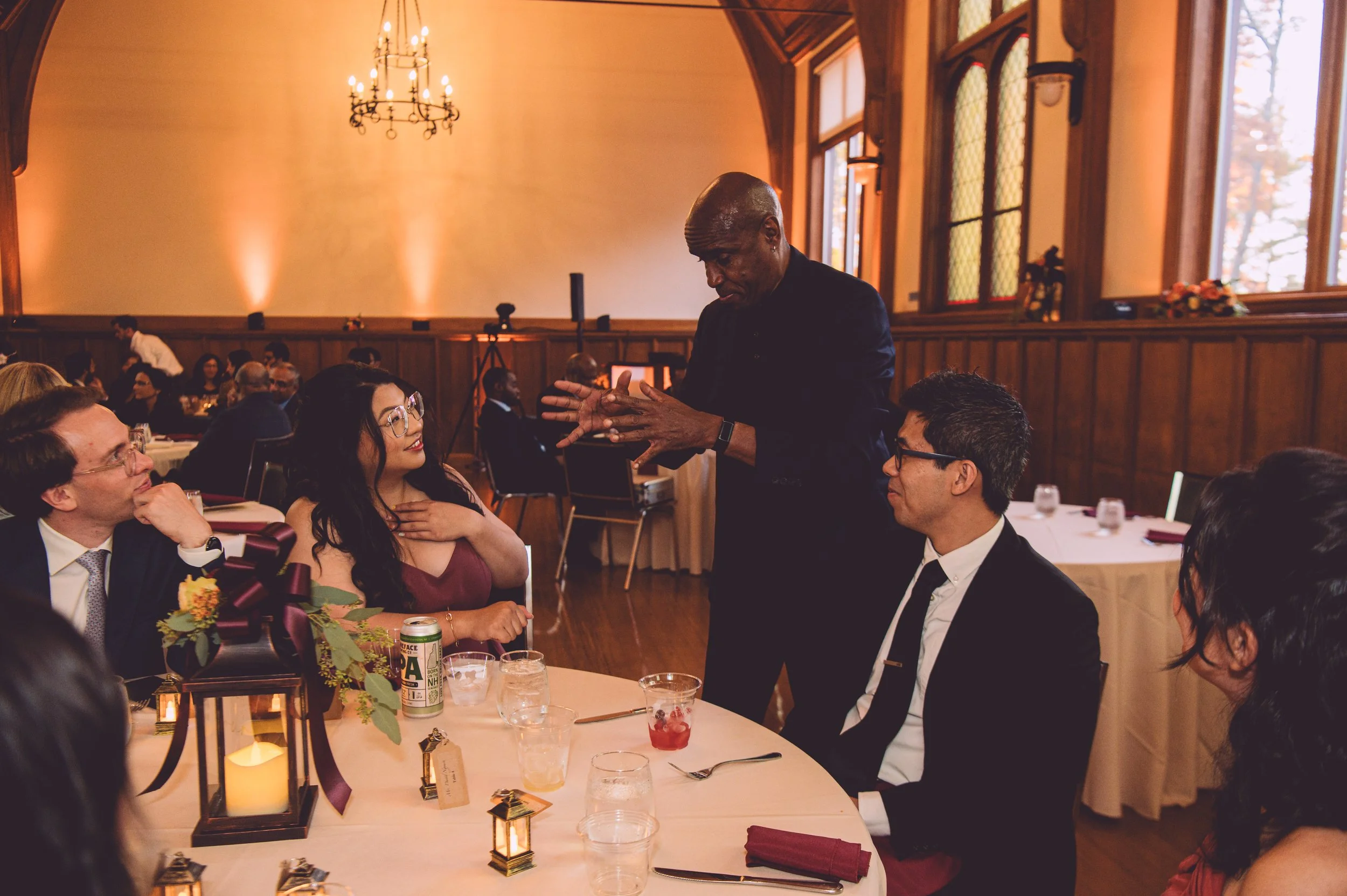 A magician performing a close-up magic trick for a table of wedding guests during a reception at Searles Chapel in Windham, NH. Wedding photography by Mike Indi.