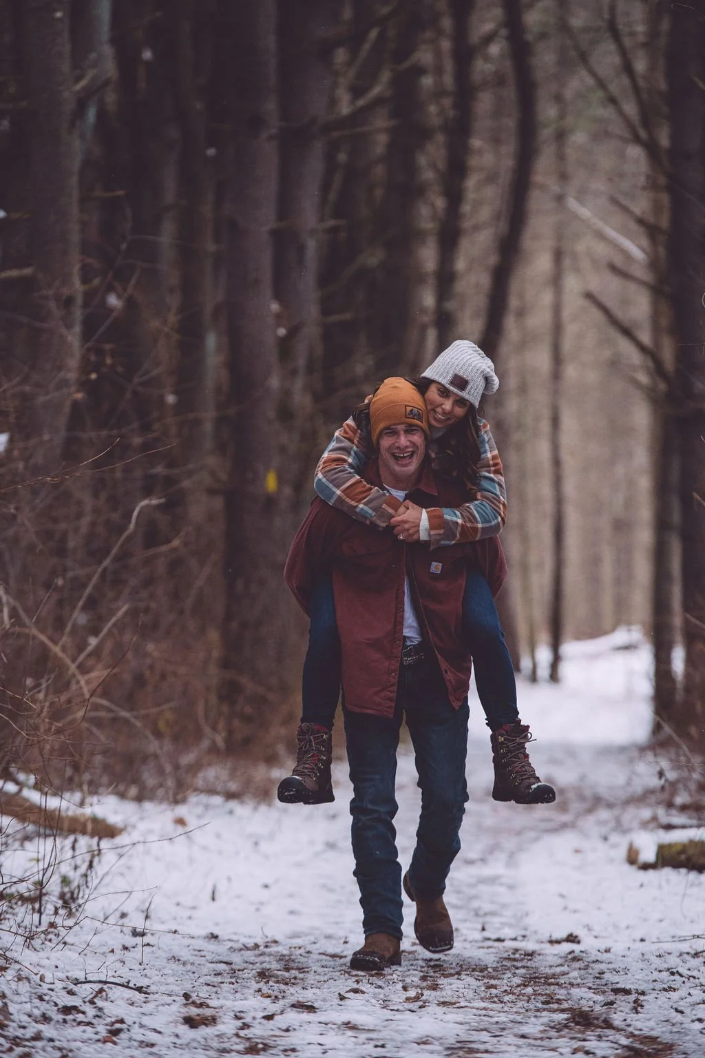 Todd is giving Ashley a piggy-back ride on a snowy wooded trail during their engagement session near Concord, NH. Photography by Mike Indi.