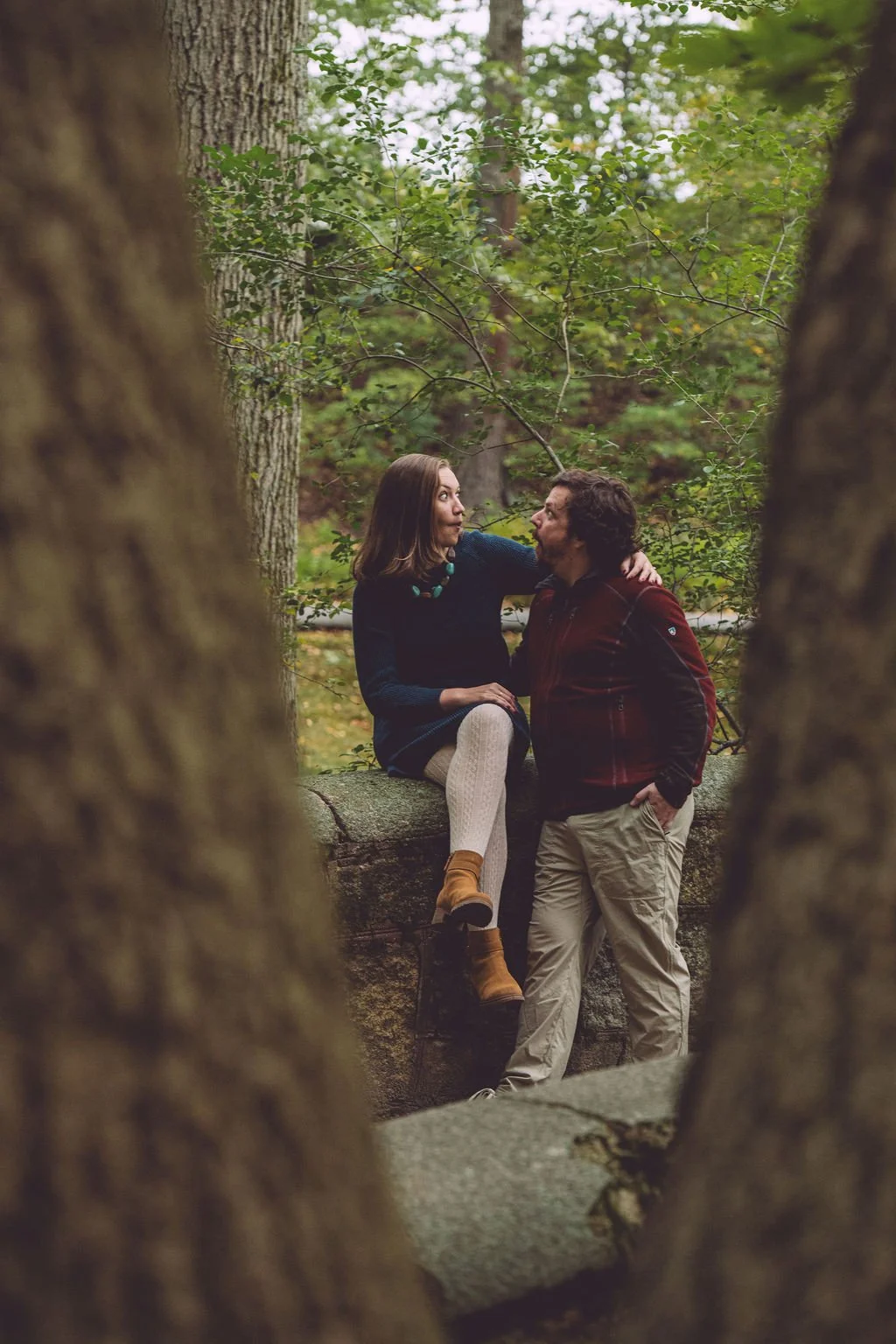 A couple making silly faces at each other on a stone bridge surrounded by lush green trees during a fall engagement session at Olmsted Park in Boston — framed through tree branches — photography by Mike Indi