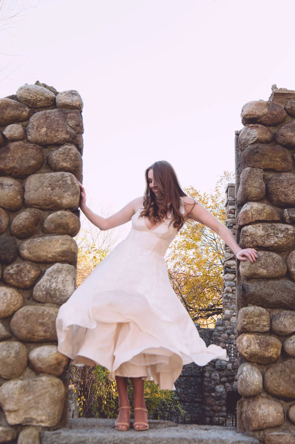 A bride dancing on stone castle ruins while the wind dramatically blows her dress during her engagement session at Bancroft's Castle in Groton, MA. Engagement photography by Mike Indi.