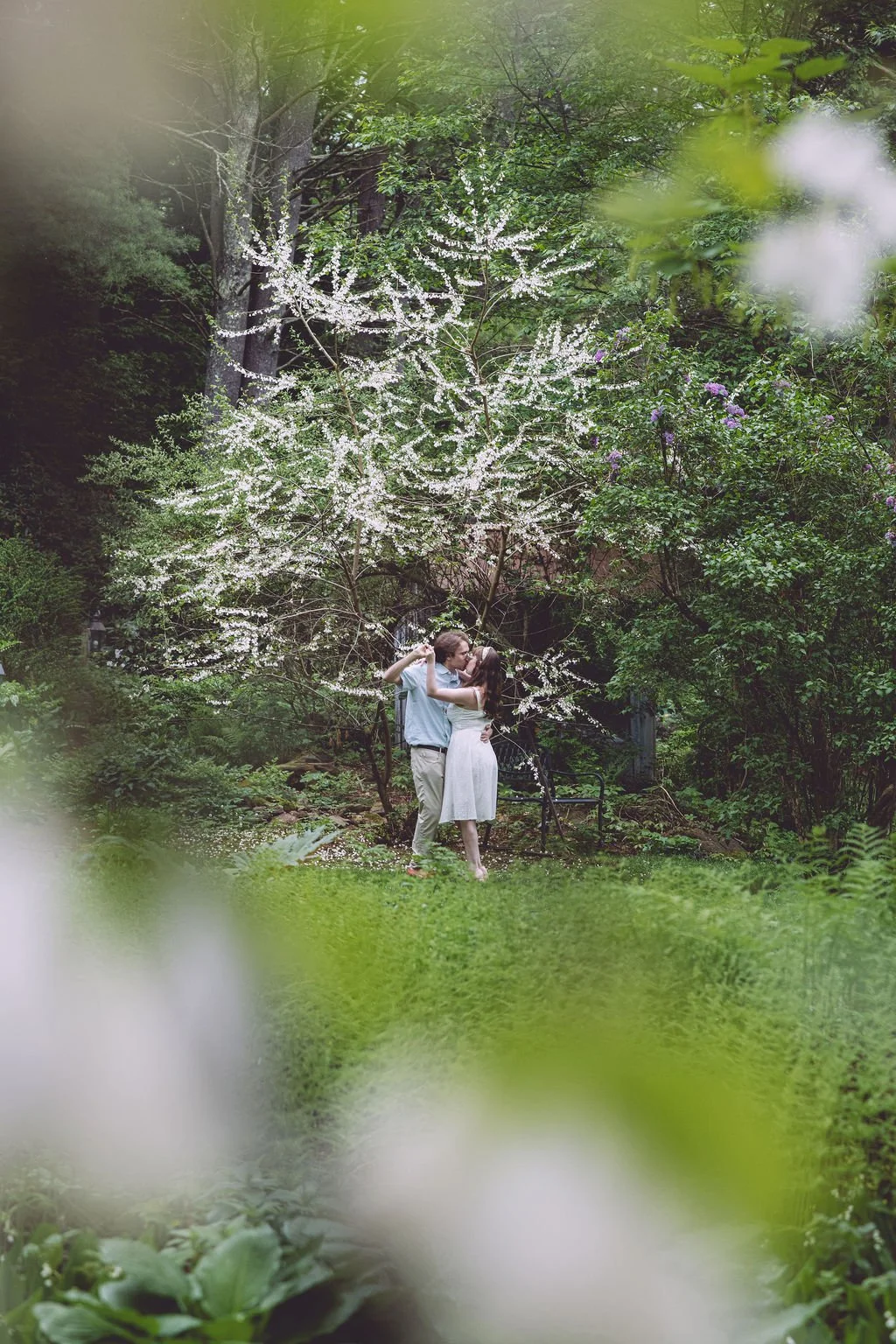 Morgan and Joey sharing a passionate kiss while dancing under a blooming tree during their spring engagement session at Tarbin Gardens in Franklin, NH. Photography by Mike Indi.