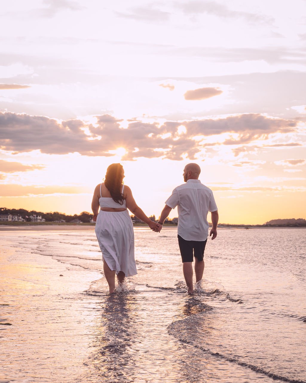 A couple wearing white holding hands and walking towards the sunset during their beach engagement session near Boston. Engagement photography by Mike Indi.