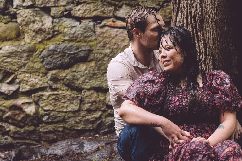 Chelsea and Ryan cuddling against a stone wall soaked from the rain during their spring engagement session at Bancroft's Castle in Groton, MA. Photography by Mike Indi.