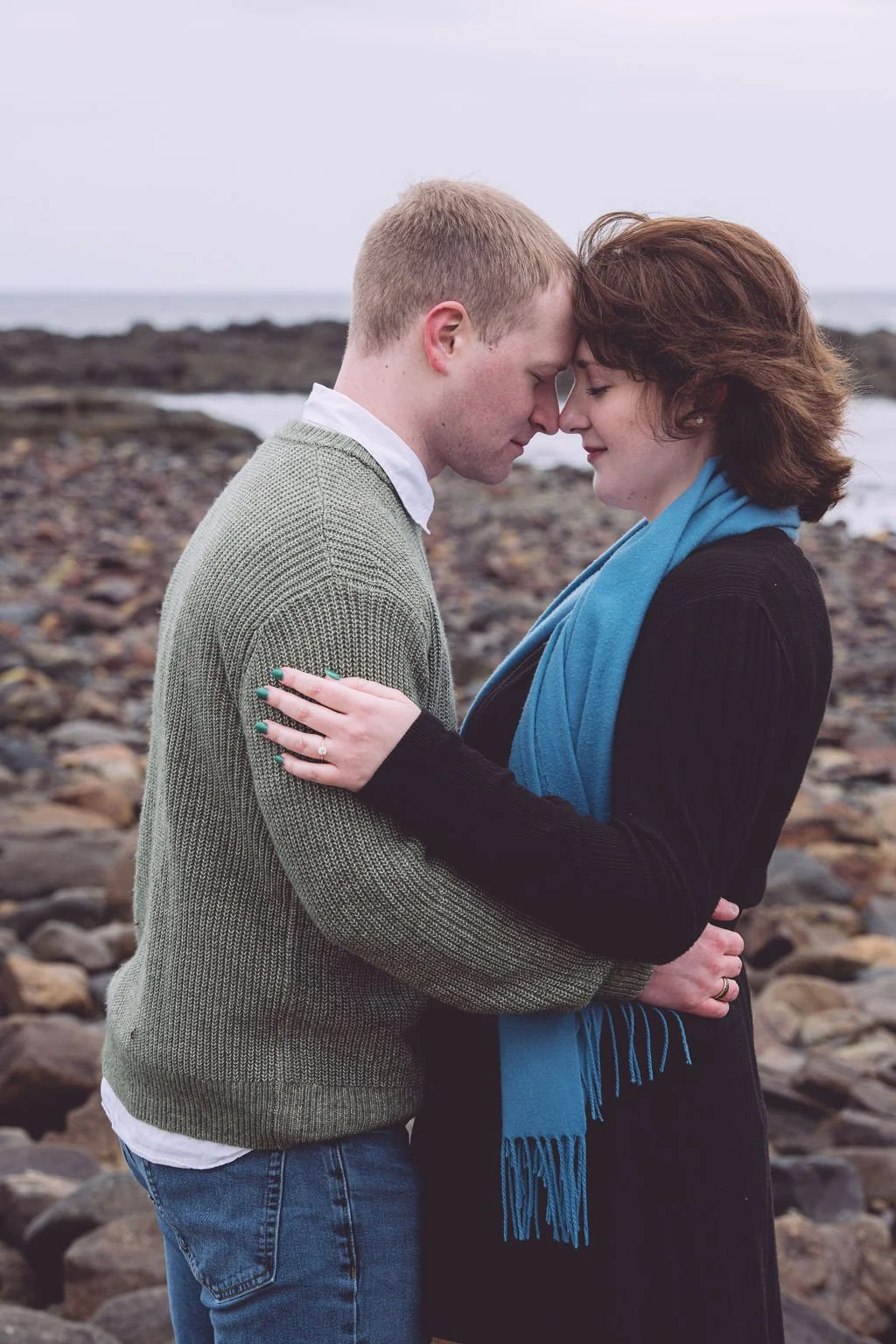 A young couple in a romantic embrace as they rest their heads on each other's and both their eyes are closed as they share a moment during their engagement session at Odiorne Point State Park in Rye, NH. Engagement photography by Mike Indi.