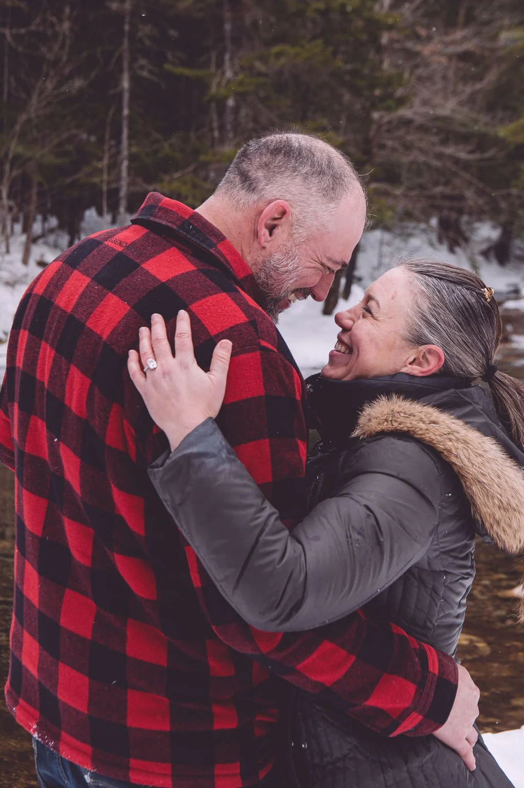 Victoria and Jason smiling while in a romantic embrace during their winter engagement session at The Basin in Franconia Notch, NH. Victoria's gorgeous ring is prominent with her hand on his shoulder. Photography by Mike Indi.