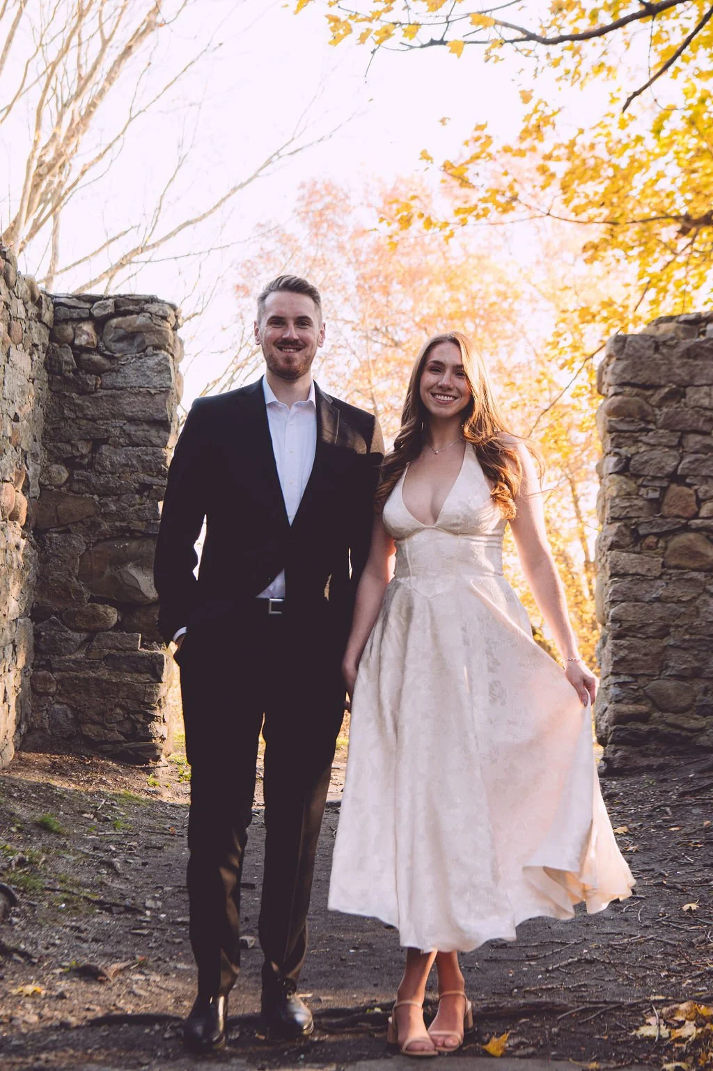 A bride and groom smiling and holding hands among the stone ruins of Bancroft's Castle in Groton, MA during their fall engagement session. Engagement photography by Mike Indi.