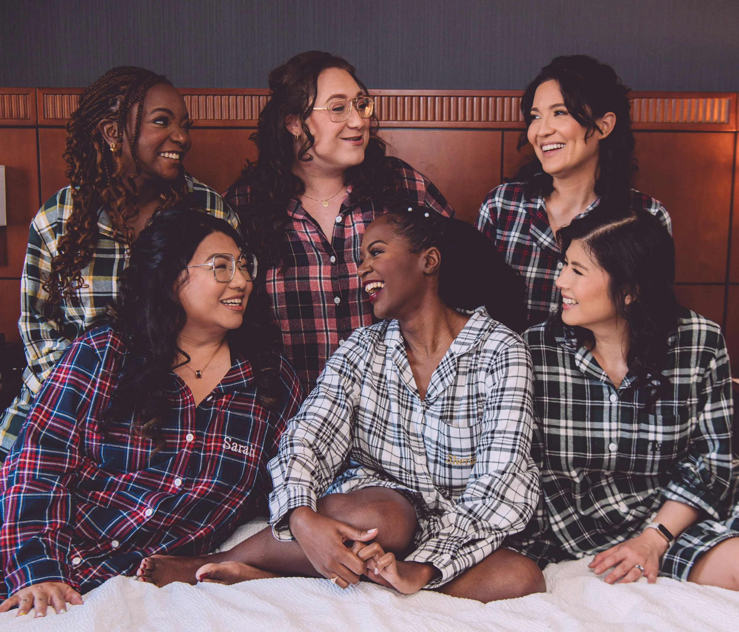 A gorgeous bride laughing with her bridesmaids while sitting on the bed in the hotel room and wearing plaid pajamas while getting ready for her wedding at Searles Chapel in Windham, NH. Wedding photography by Mike Indi.