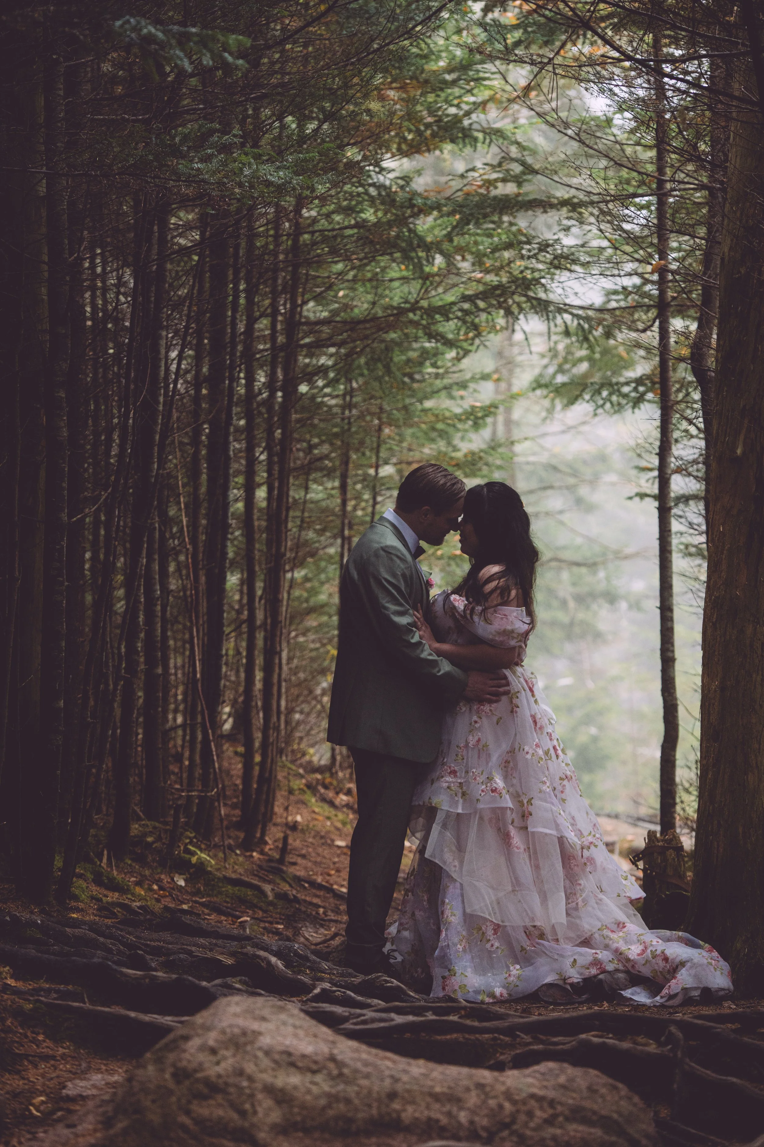 Chelsea and Ryan sharing a romantic kiss surrounded by trees and natural light coming through on a foggy morning during their adventure elopement in Acadia National Park in Bar Harbor, Maine - Photography by Mike Indi